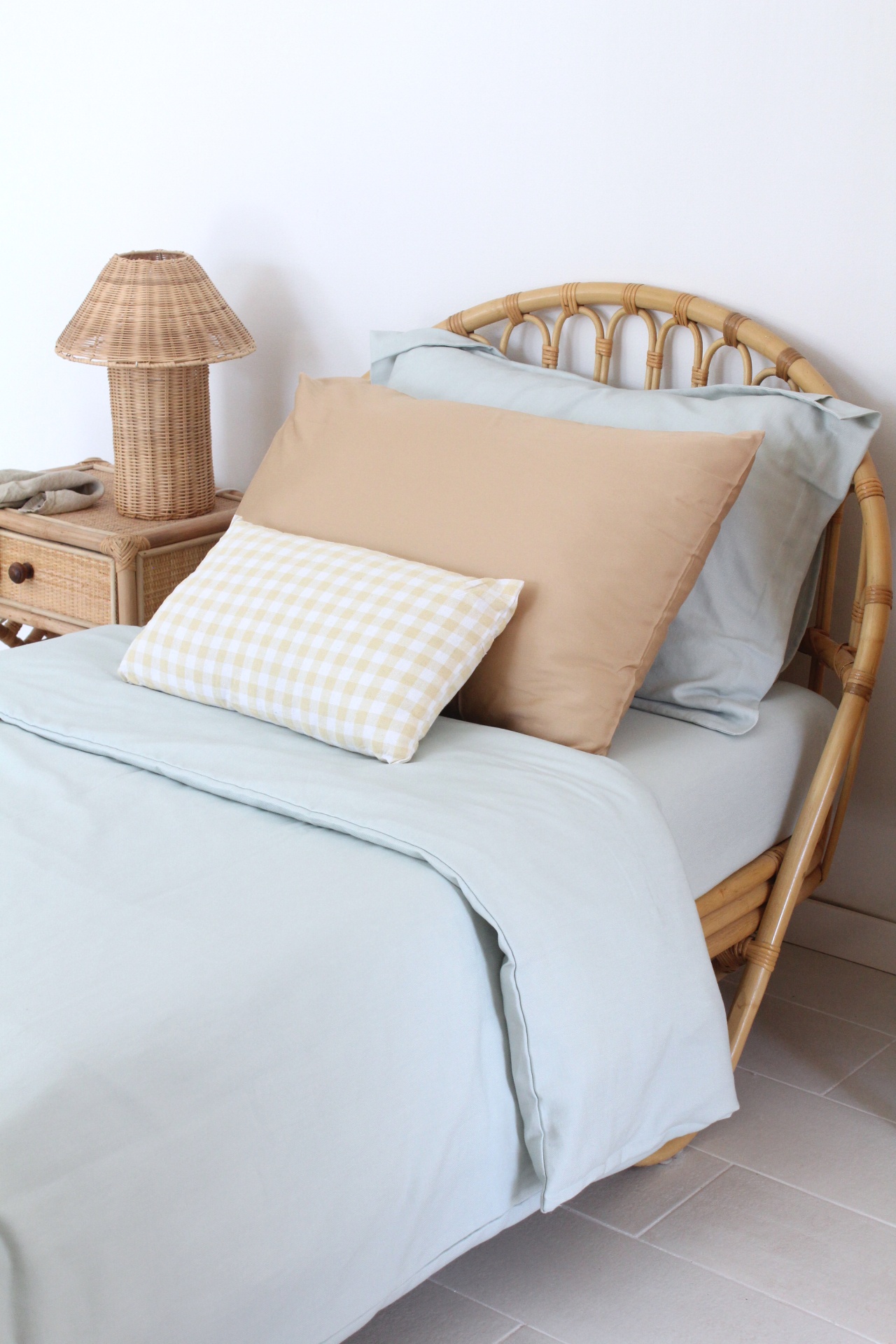 Cama com estrutura em rattan, roupa de cama azul claro, almofadas bege e xadrez amarelo e branco, e candeeiro em rattan numa mesa de cabeceira