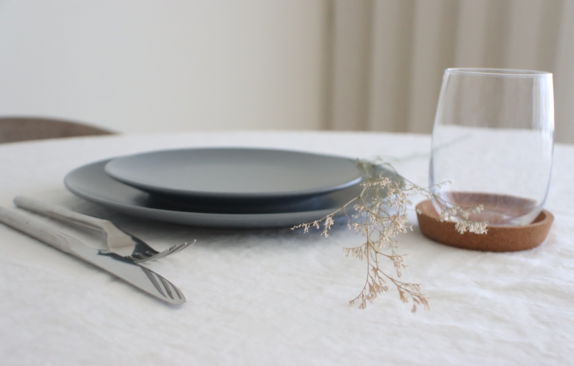 Conjunto de prato cinzento, talheres metálicos e copo com base em cortiça numa mesa com toalha branca