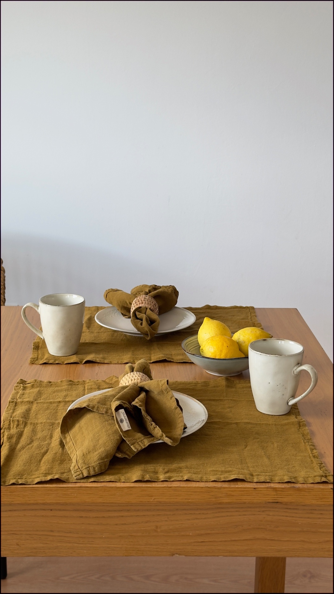 Mesa de madeira com dois jogos americanos castanhos, pratos brancos, guardanapos castanhos com argolas de corda, copos brancos e tigela com limões amarelos.
