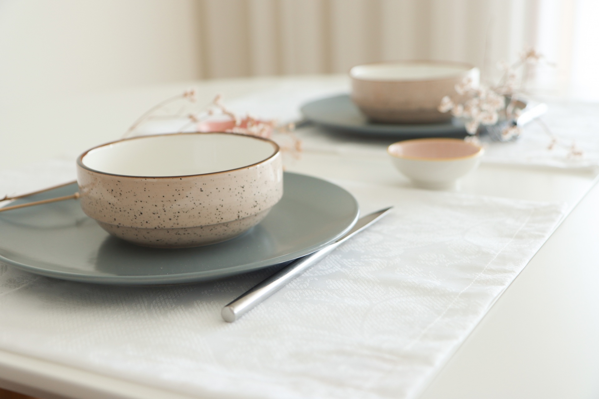 Conjunto de loiça em cerâmica bege com salpicos e prato cinza numa mesa com toalha branca.