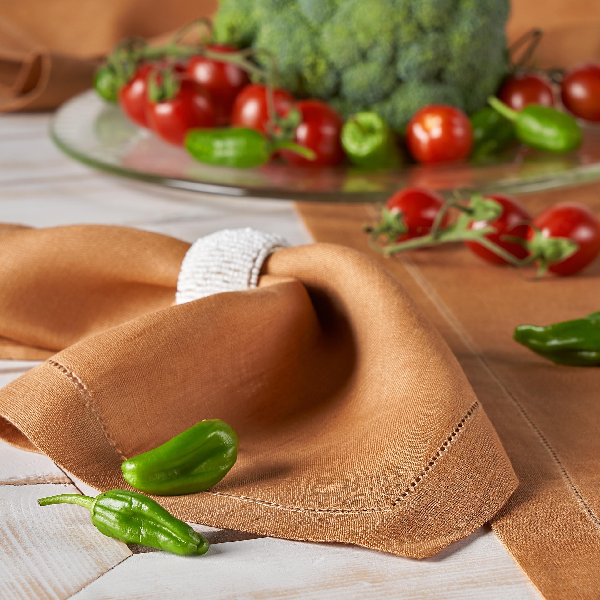 Guardanapo castanho com anel branco ao lado de malaguetas verdes e prato com tomates cereja e brócolos