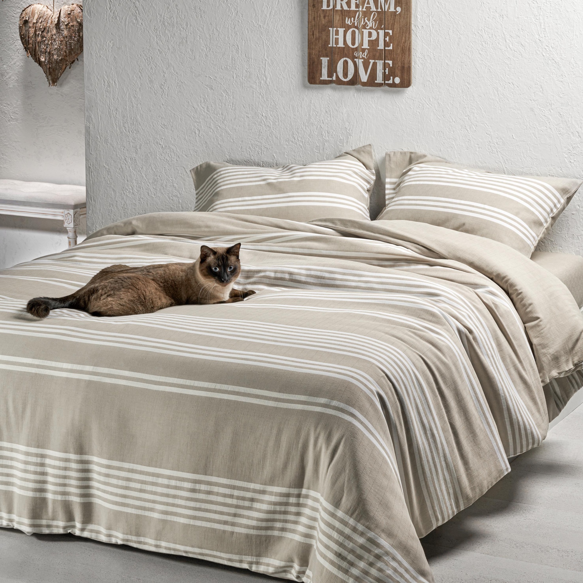Cama com roupa de cama bege às riscas, gato deitado, parede branca com placa de madeira decorativa.