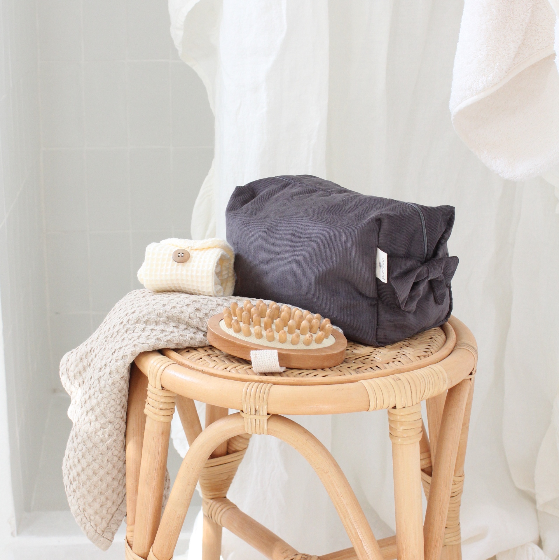 Taborete em vime claro com almofada preta, toalha bege, escova de madeira e esponja de banho, fundo branco