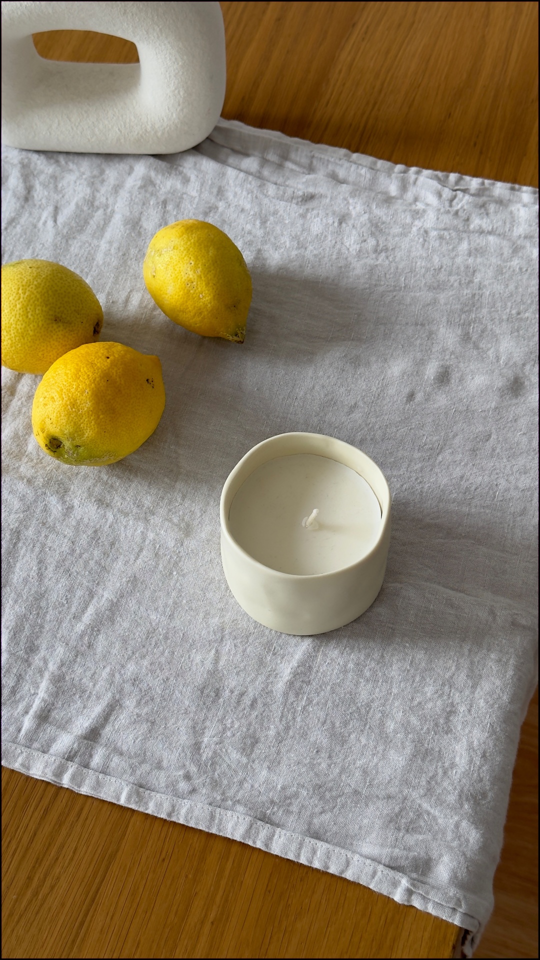 Vela branca numa base cerâmica com limões e pano cinzento sobre mesa de madeira