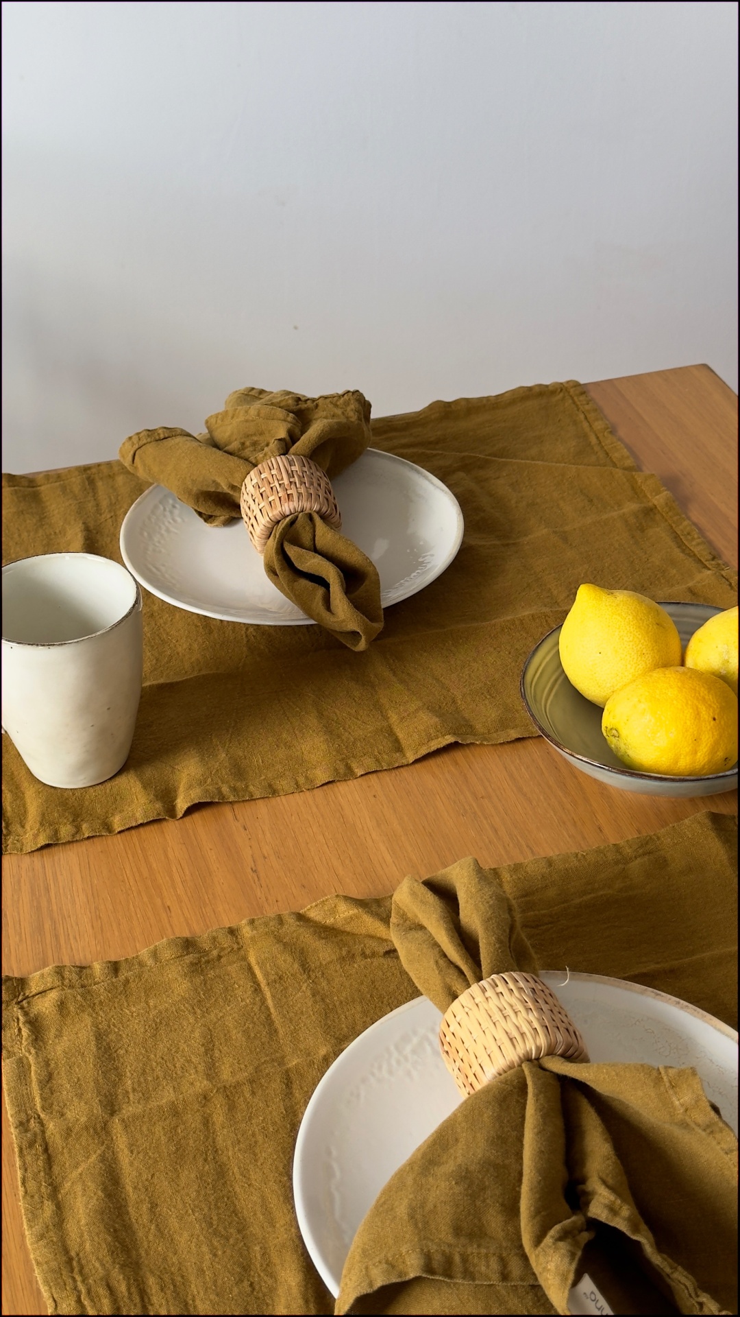 Mesa de madeira com pratos brancos, guardanapos castanhos em argolas de palha, copo branco e tigela de vidro com limões amarelos.