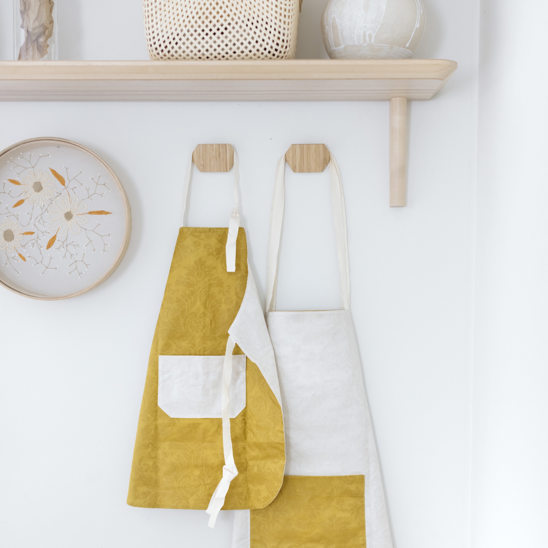 Dois aventais pendurados numa parede branca abaixo de prateleira de madeira e ao lado de quadro decorativo redondo