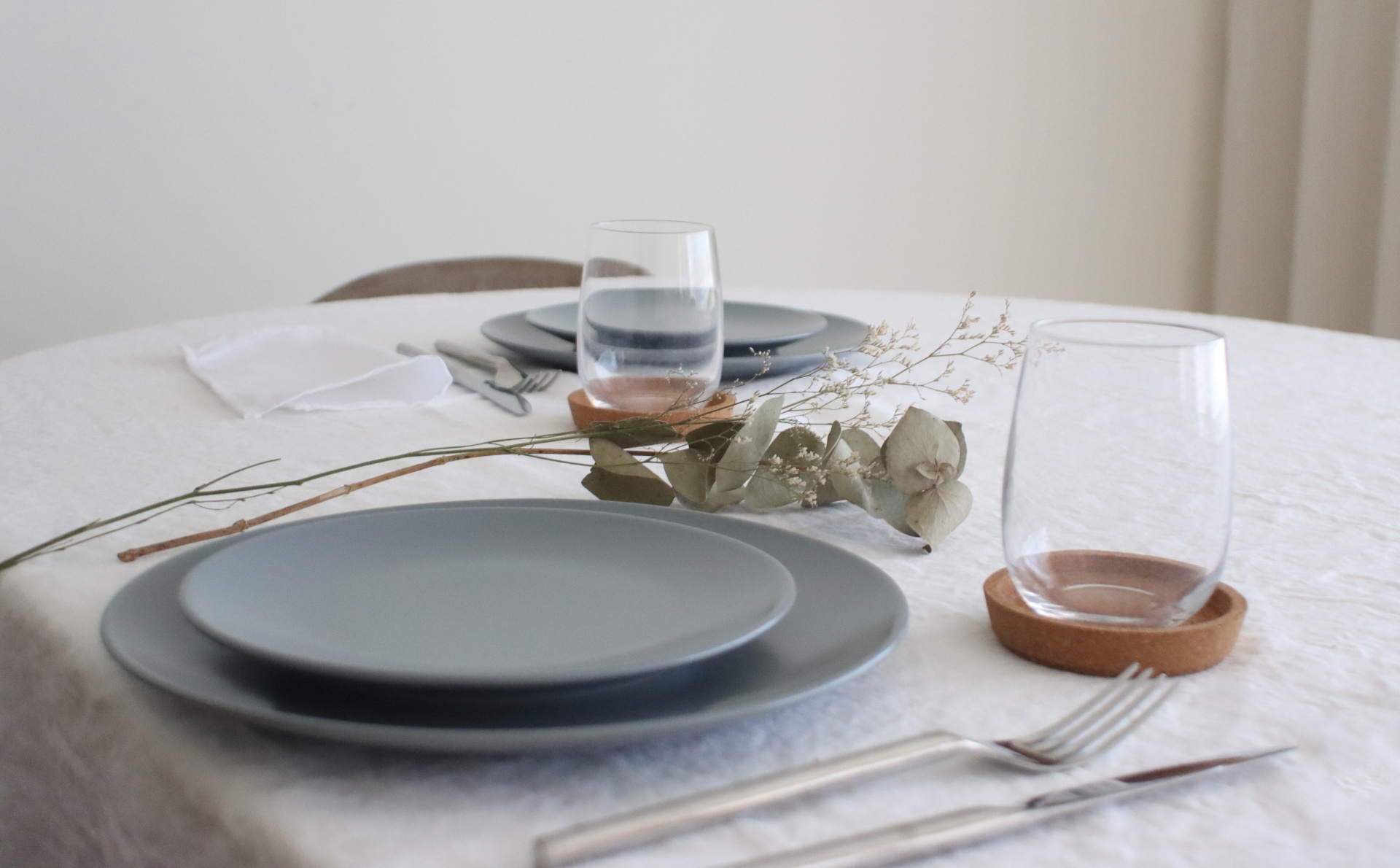 Mesa posta com pratos cinza, copos transparentes com base castanha, talheres e decoração com ramos secos