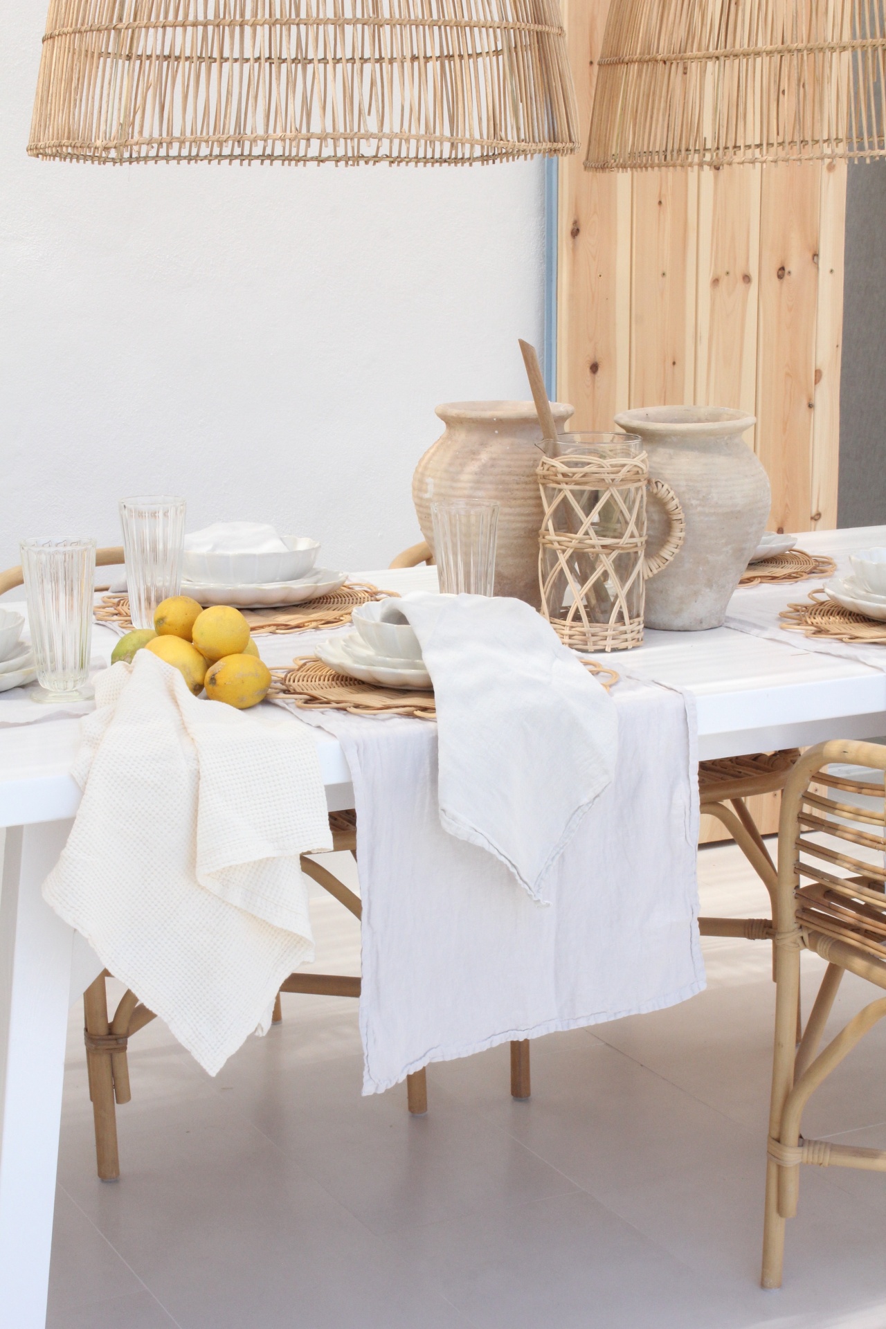 Mesa branca posta com toalhas de linho, frutas amarelas, vasos de cerâmica e abajures de vime em ambiente branco e madeira clara