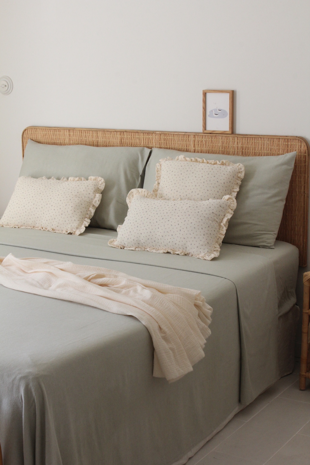 Cama com roupa de cama verde-acinzentada e cabeceira em rattan com almofadas creme e manta