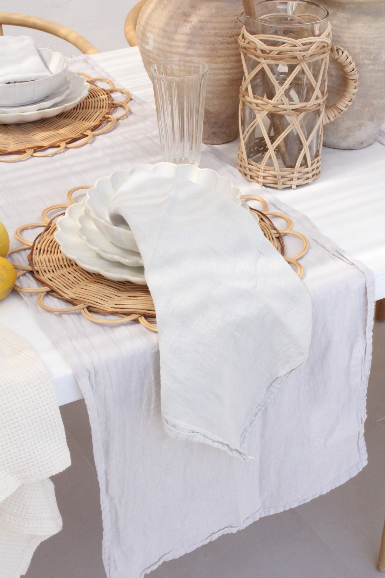 Mesa posta com pratos brancos, guardanapo, copo, jarra com suporte de rattan e jarros de cerâmica