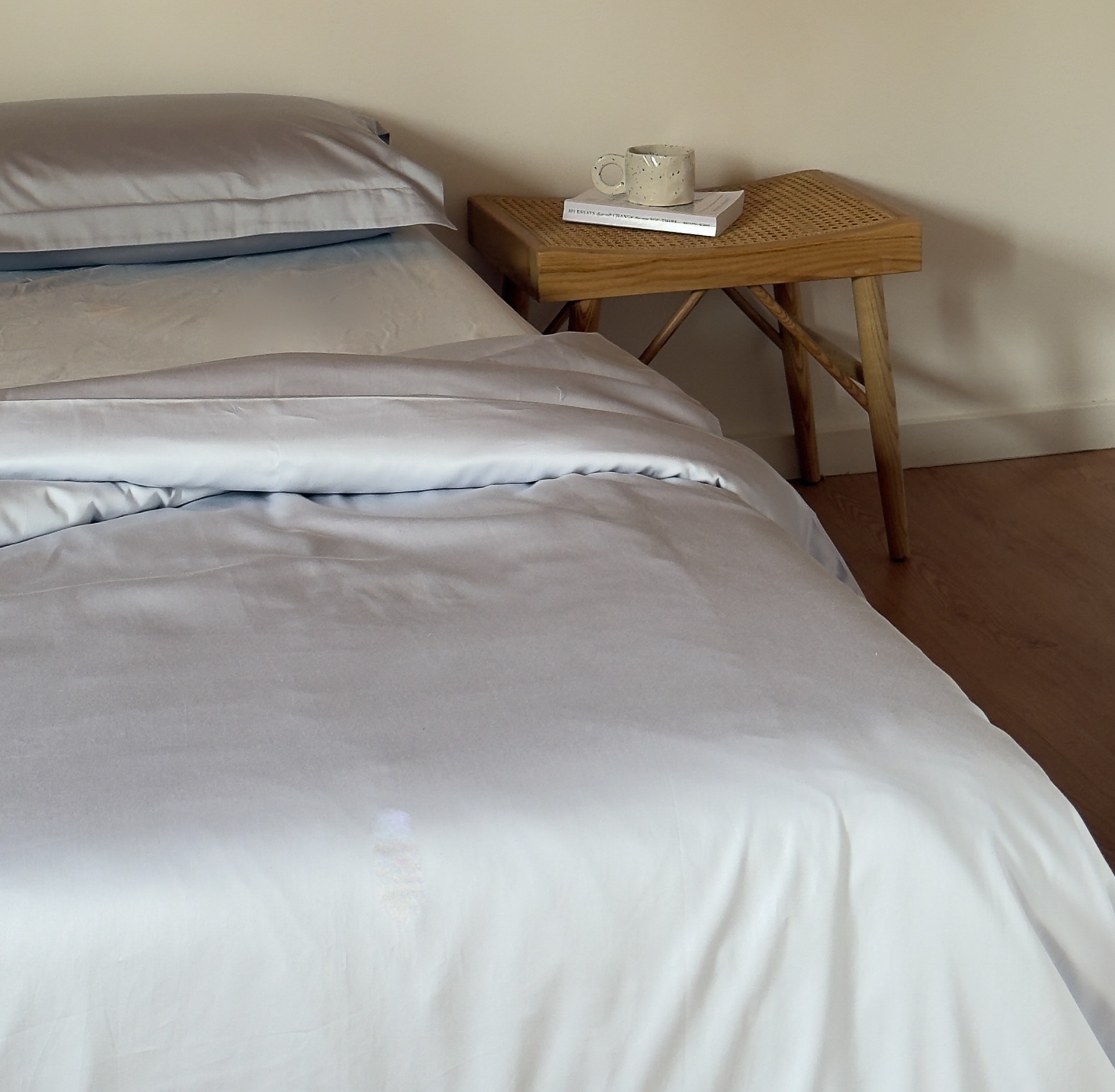 Cama com lençol bege claro e mesa de apoio de madeira com copo e livro