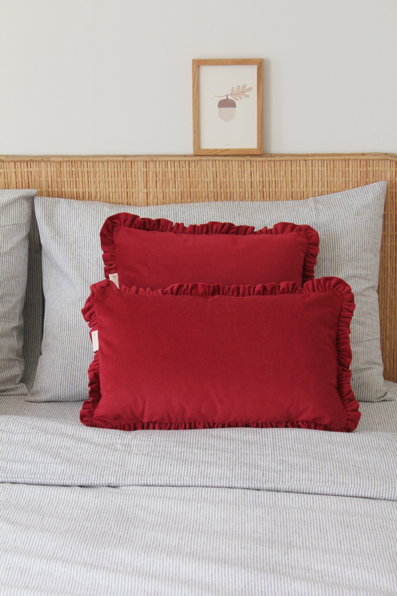 Duas almofadas vermelhas com folhos numa cama com roupa de cama cinzenta riscada e cabeceira em rattan.