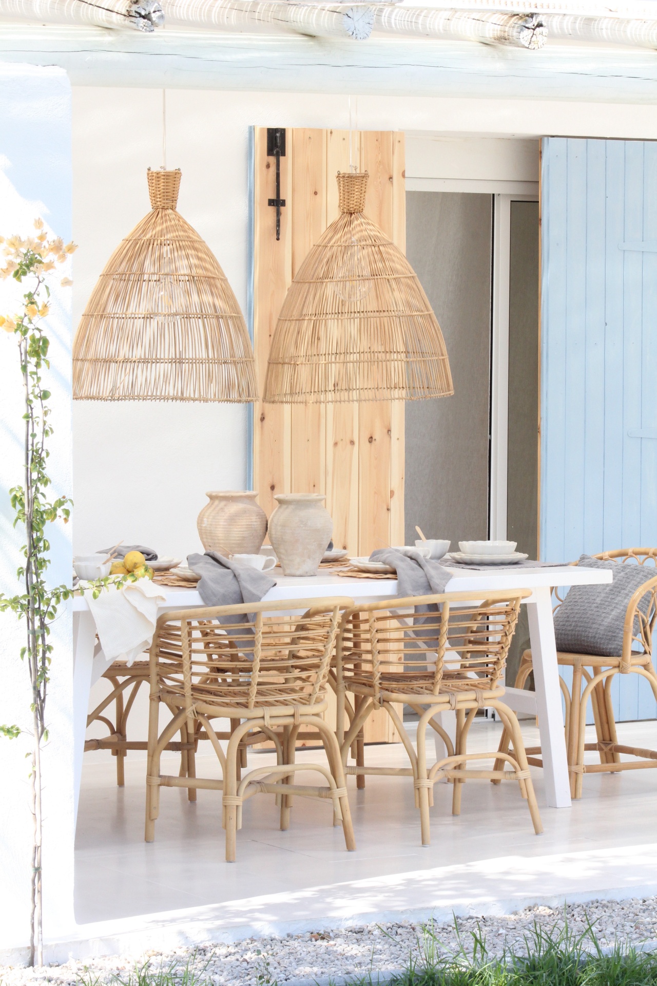 Mesa branca retangular com cadeiras e candeeiros suspensos em rattan em espaço exterior com paredes brancas e persianas azuis.