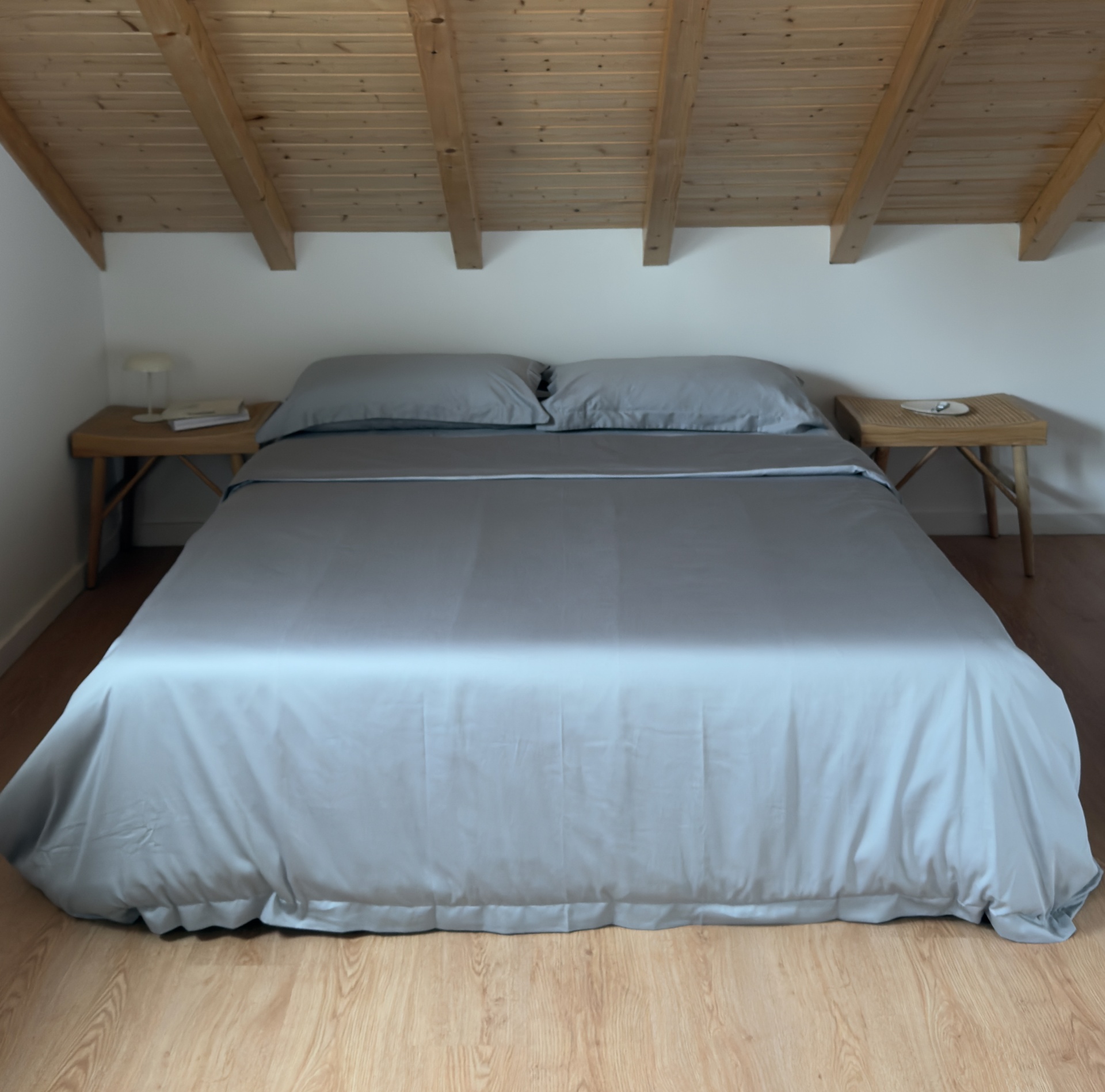 Cama de casal com lençóis cinza, mesas de cabeceira e tecto em madeira inclinado