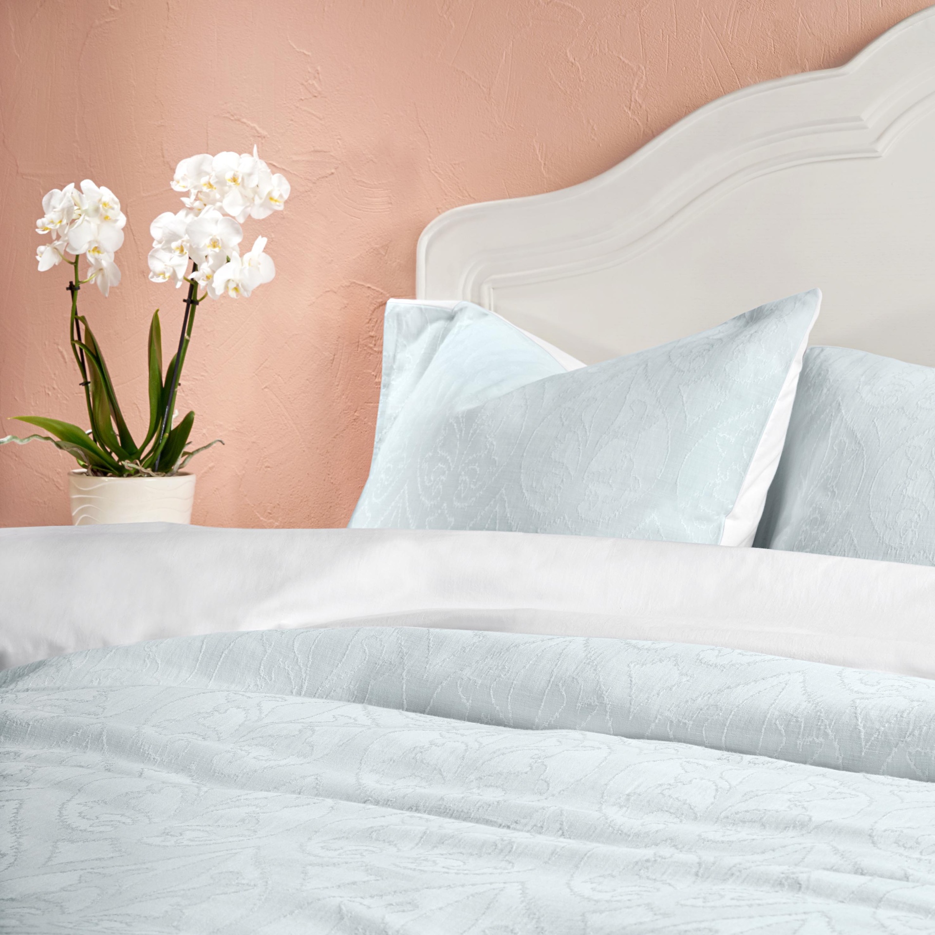 Conjunto de roupa de cama azul claro com padrão sutil, cama branca, orquídeas brancas e parede salmão