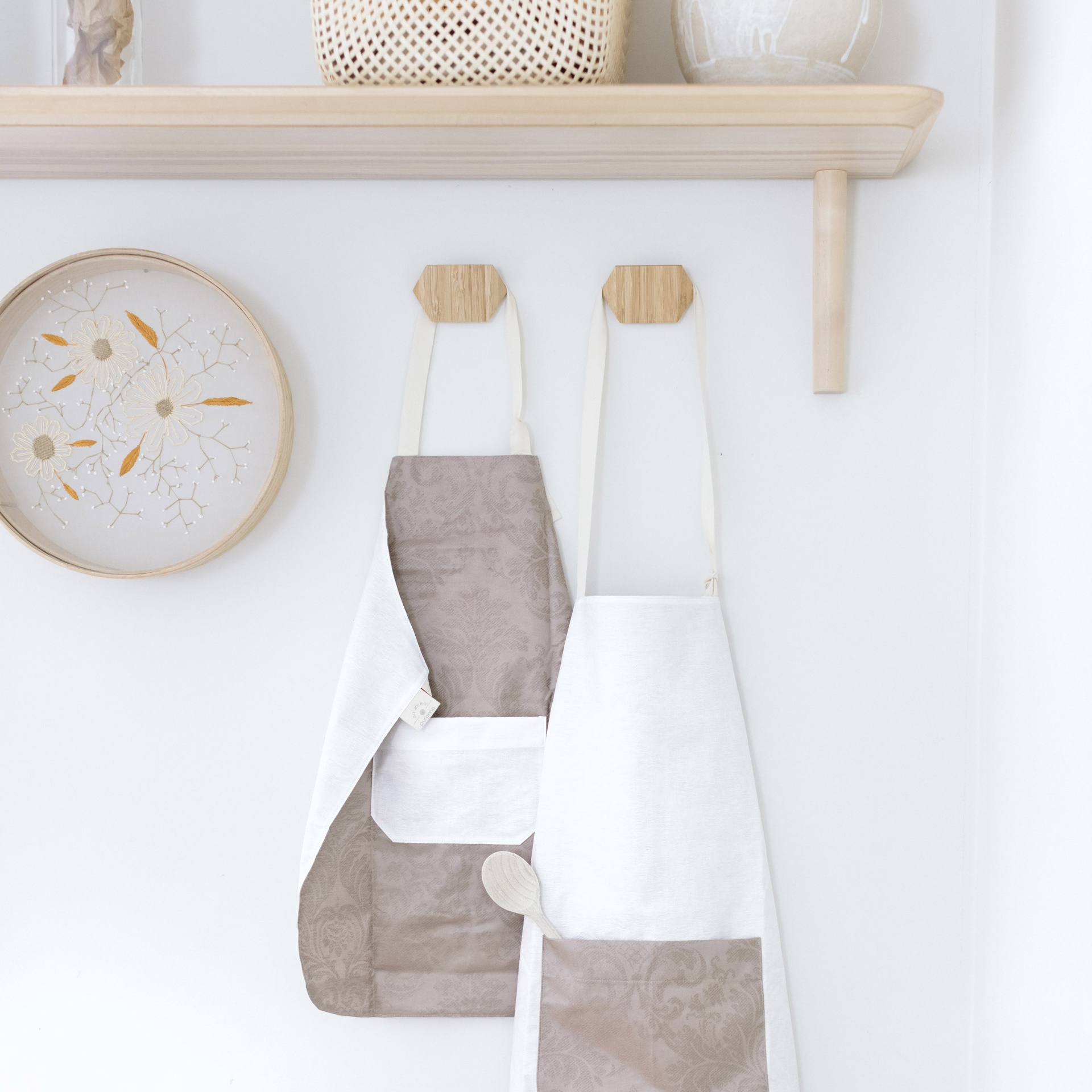 Dois aventais bege e branco pendurados numa parede branca com relógio decorativo e prateleira de madeira