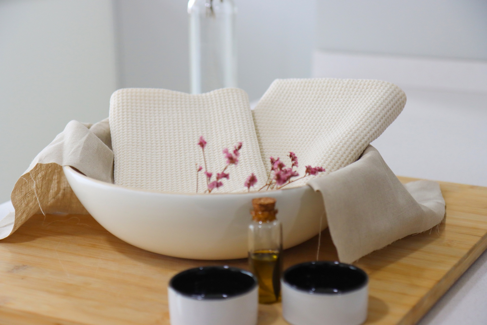 Toalhas creme de tecido tipo waffle em taça branca com flores secas, duas pequenas taças e frasco de vidro com líquido