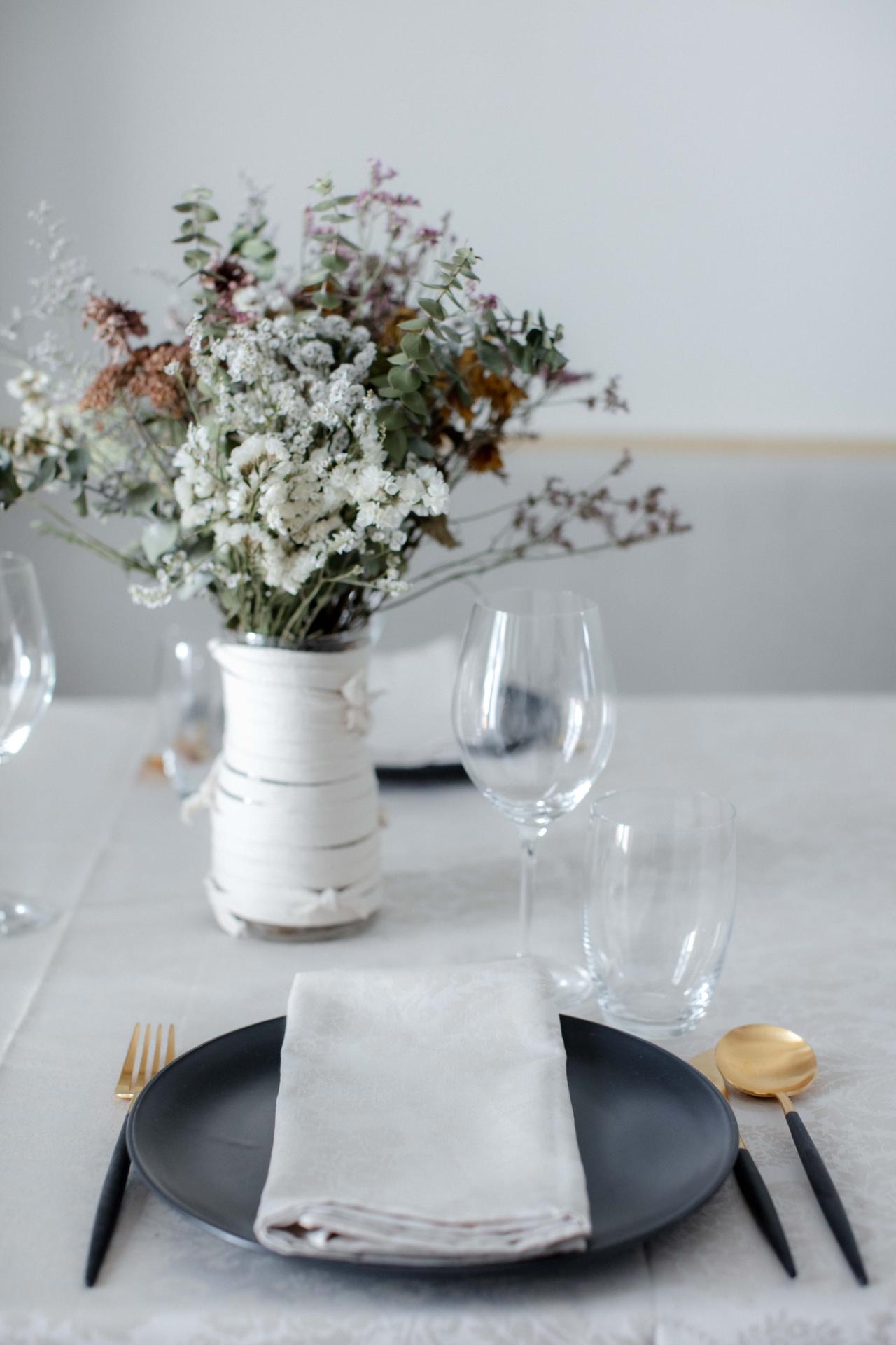 Conjunto de jantar com prato preto, guardanapo branco, talheres dourados e pretos, copos e vaso com flores secas
