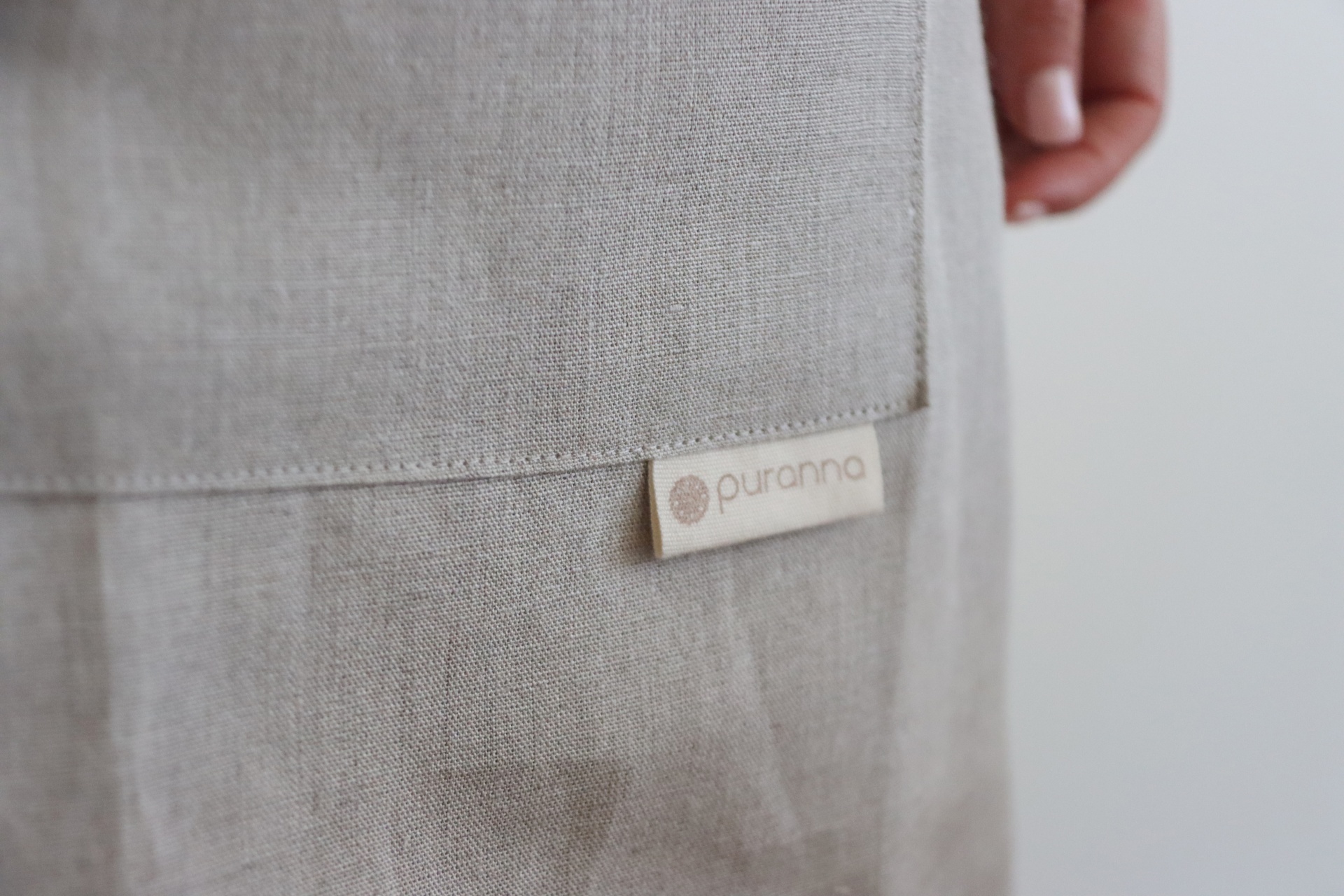 Etiqueta de tecido com 'puranna' em roupa cinza claro com textura e mão ao fundo