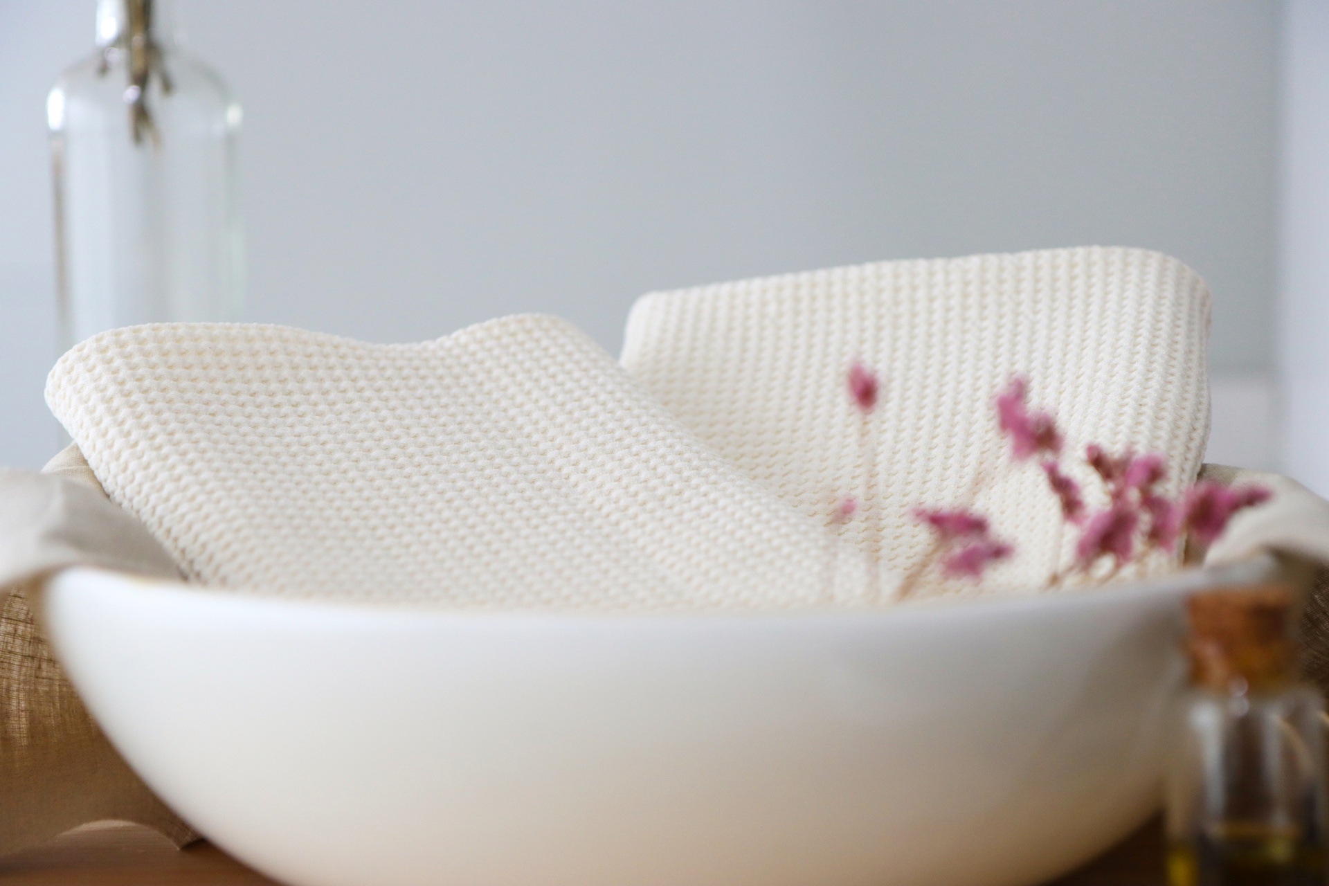 Toalhas brancas texturizadas numa bacia branca com flores cor-de-rosa e frasco ao fundo