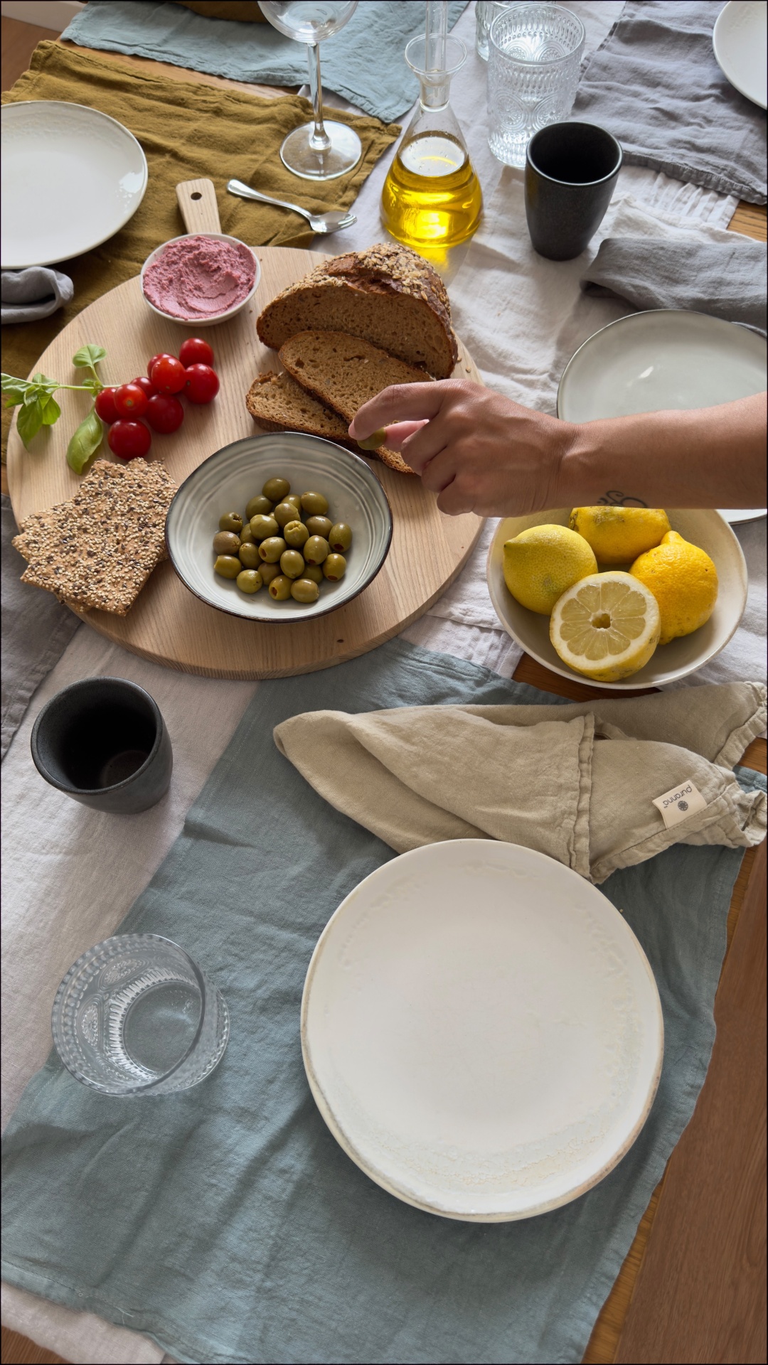 Mesa posta com pão, azeitonas, tomates, limões e azeite sobre tábua redonda de madeira, pratos e copos sobre toalhas de mesa coloridas.