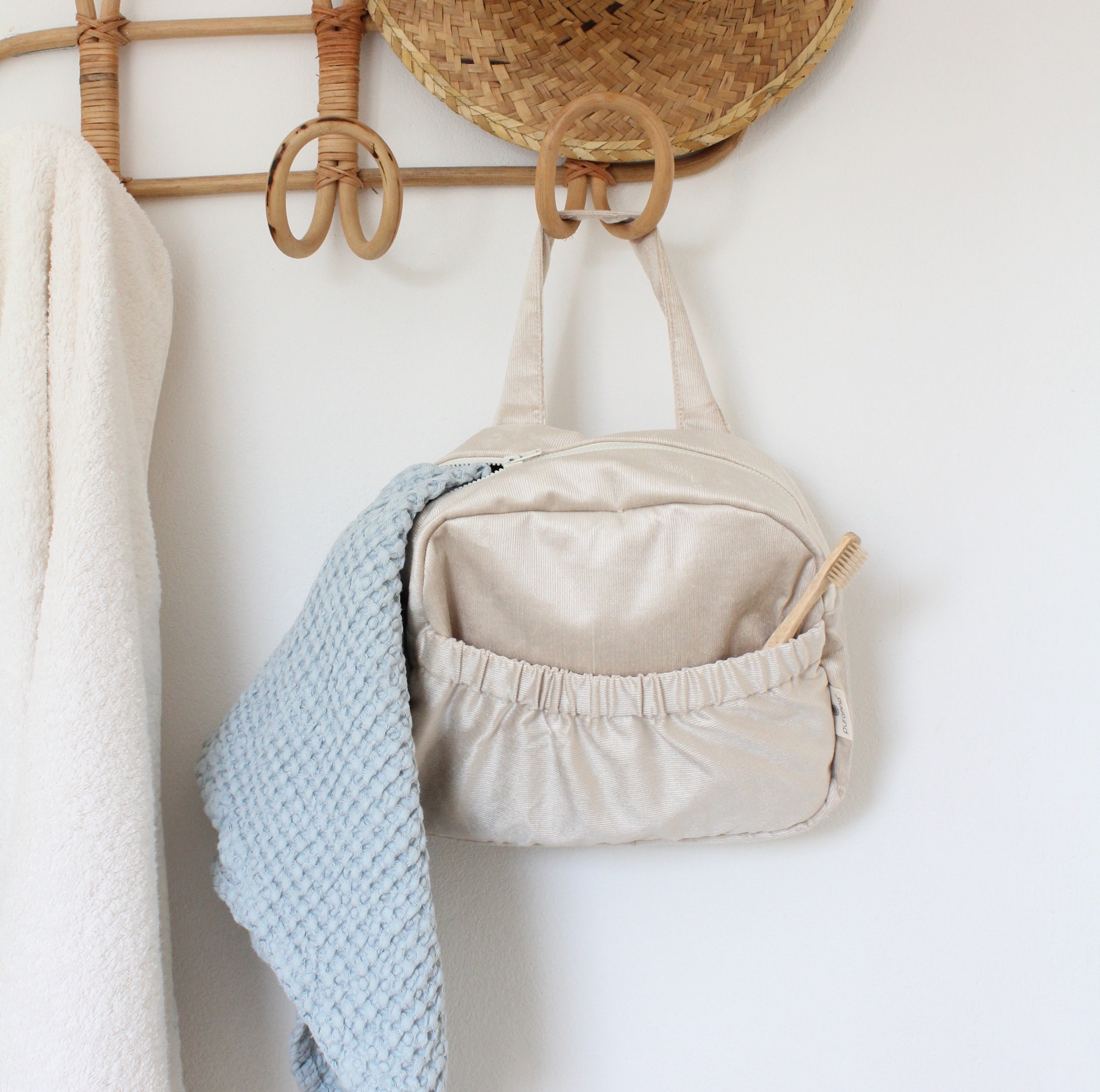 Mala de tecido bege pendurada num cabide de madeira com toalha azul e escova, fundo branco e chapéu de palha.
