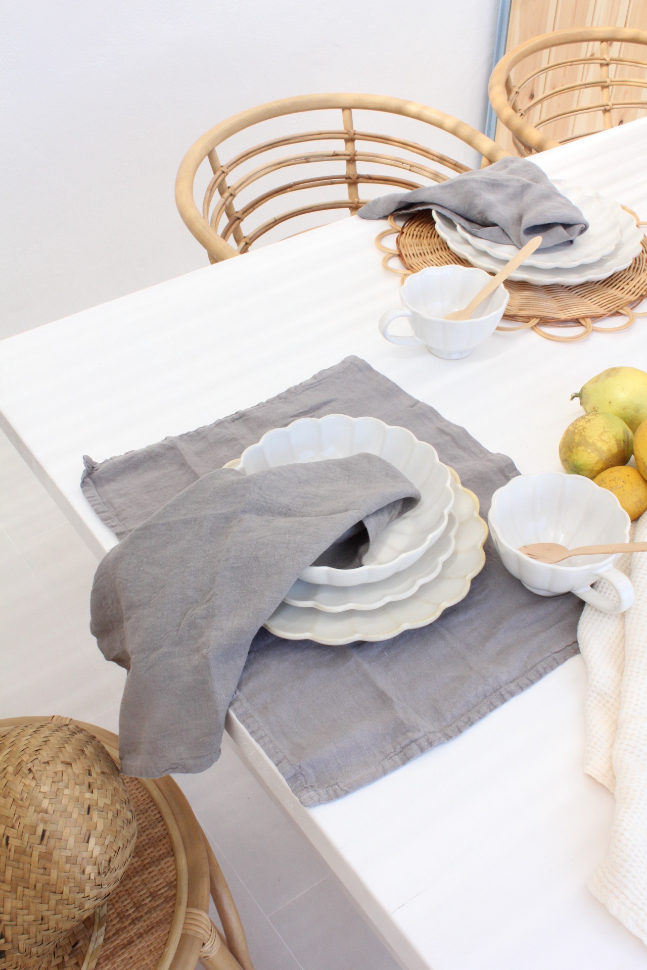 Mesa branca com conjunto de loiça branca ondulada, panos cinzentos e cadeiras de rattan