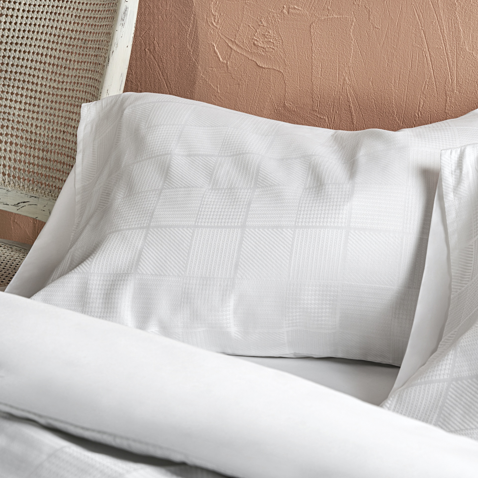 Almofada branca com fronhas em tecido texturizado quadrado e lençol branco numa cama junto a parede cor salmão