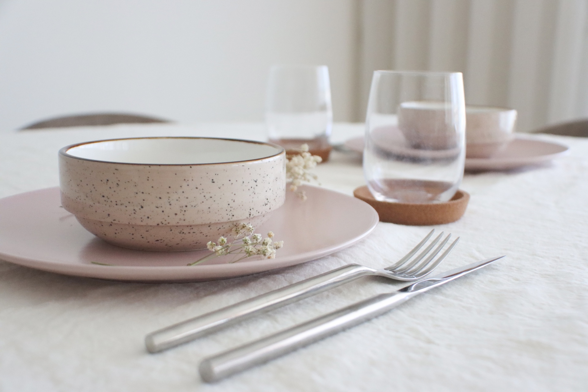 conjunto de loiça bege e rosa claro com copos de vidro e talheres numa mesa com toalha branca