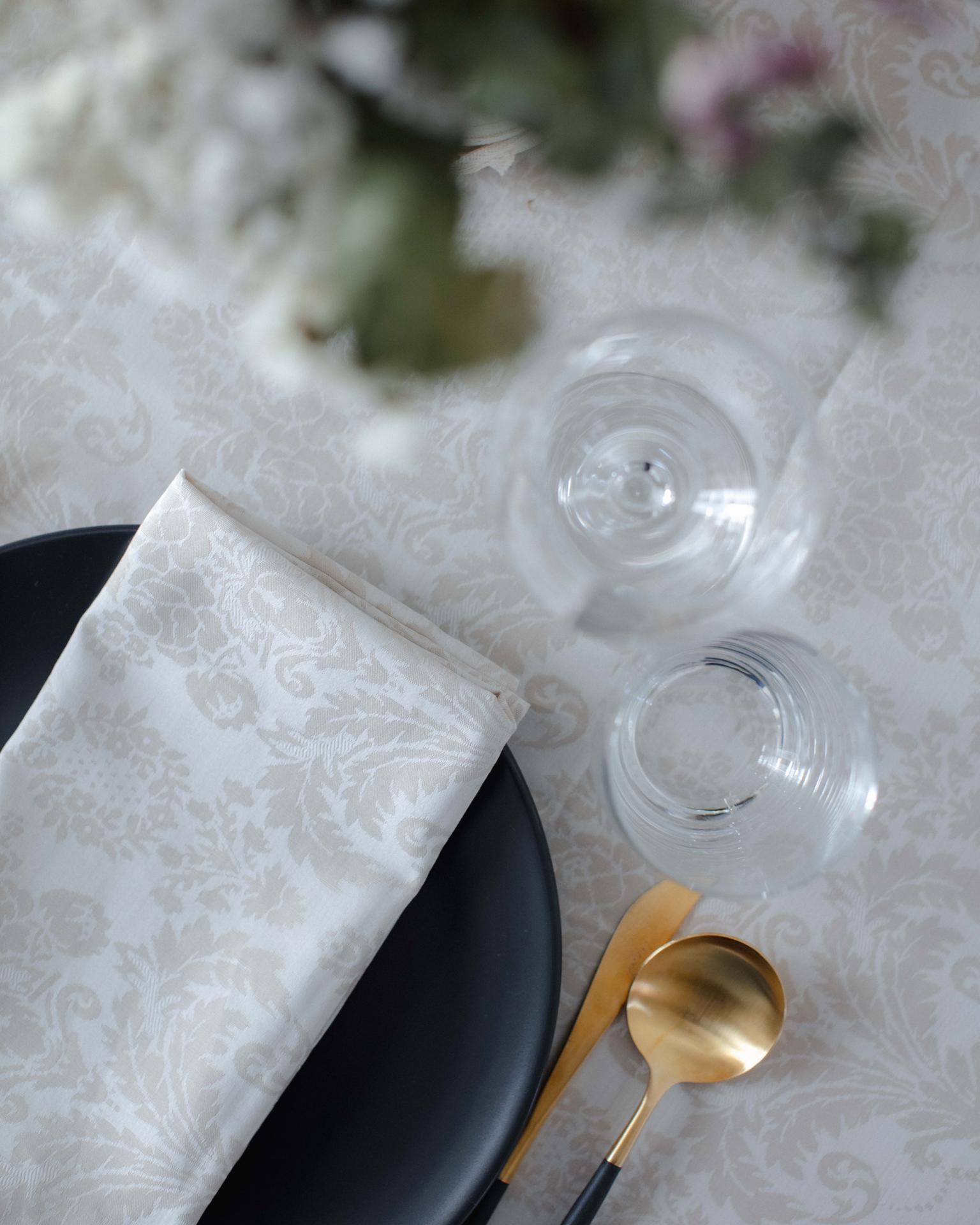 Mesa com toalha branca floral, prato preto, guardanapo branco, talheres dourados e copos transparentes