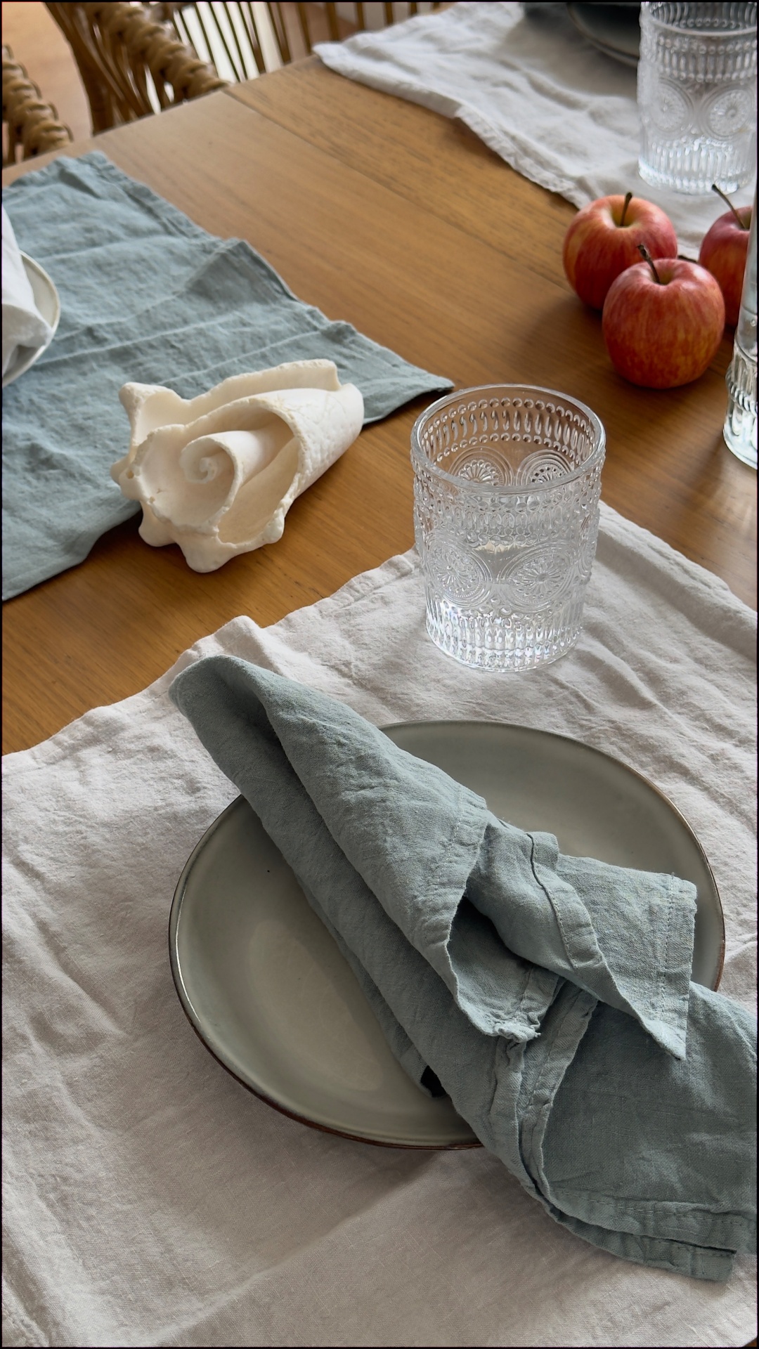 Mesa posta com prato cinza, guardanapo azul, copo decorado, maçãs e concha