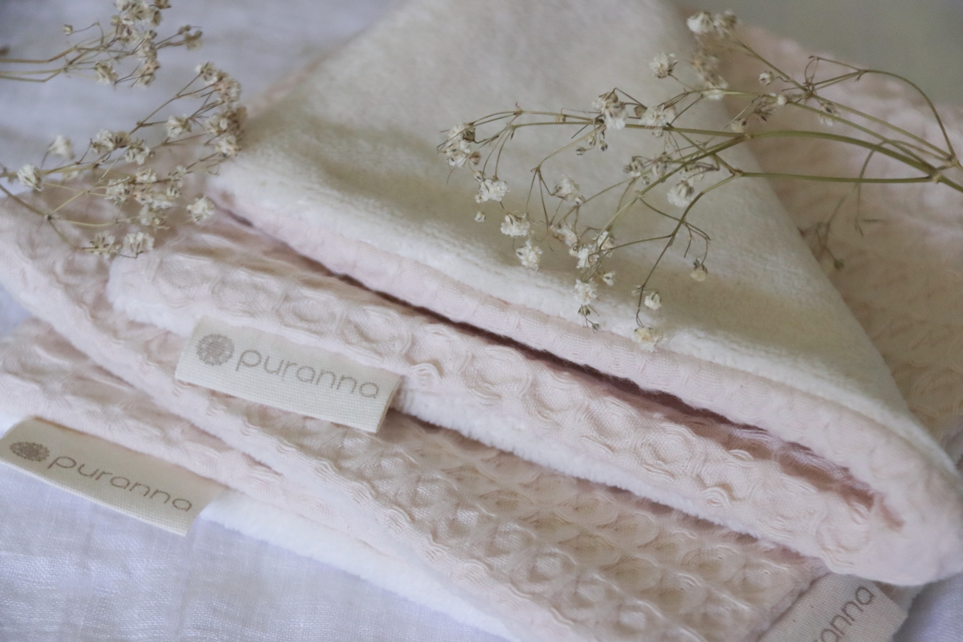 Toalhas de banho cor-de-rosa e branca com etiquetas 'puranna' e flores secas em cima