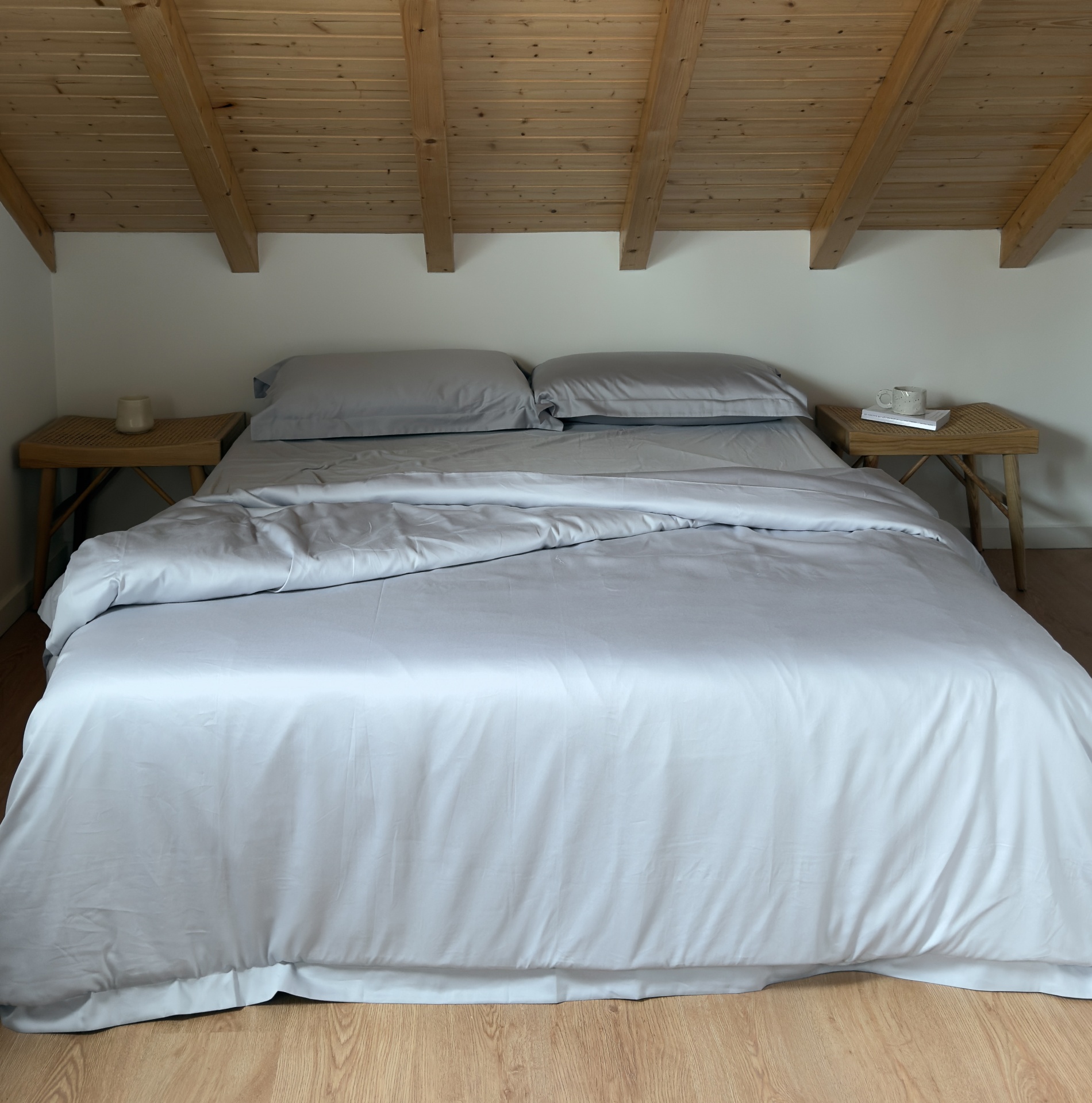 Cama de casal com roupa de cama cinza clara sob teto inclinado de madeira