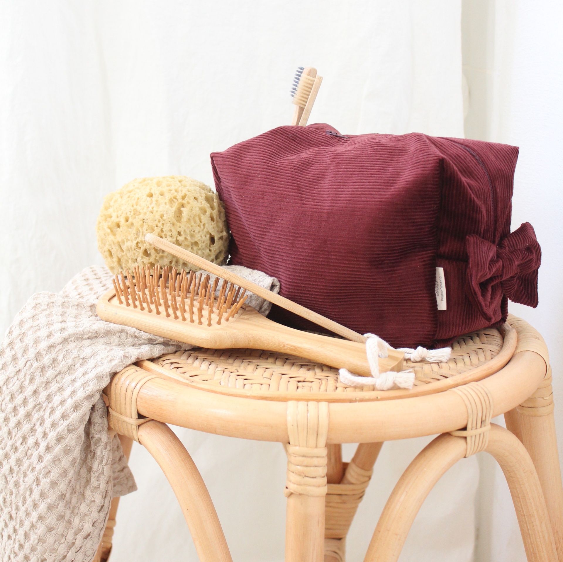 Acessórios de higiene pessoal em banco de rattan com necessaire vermelha, escova e pente de madeira, esponja natural e toalha bege.