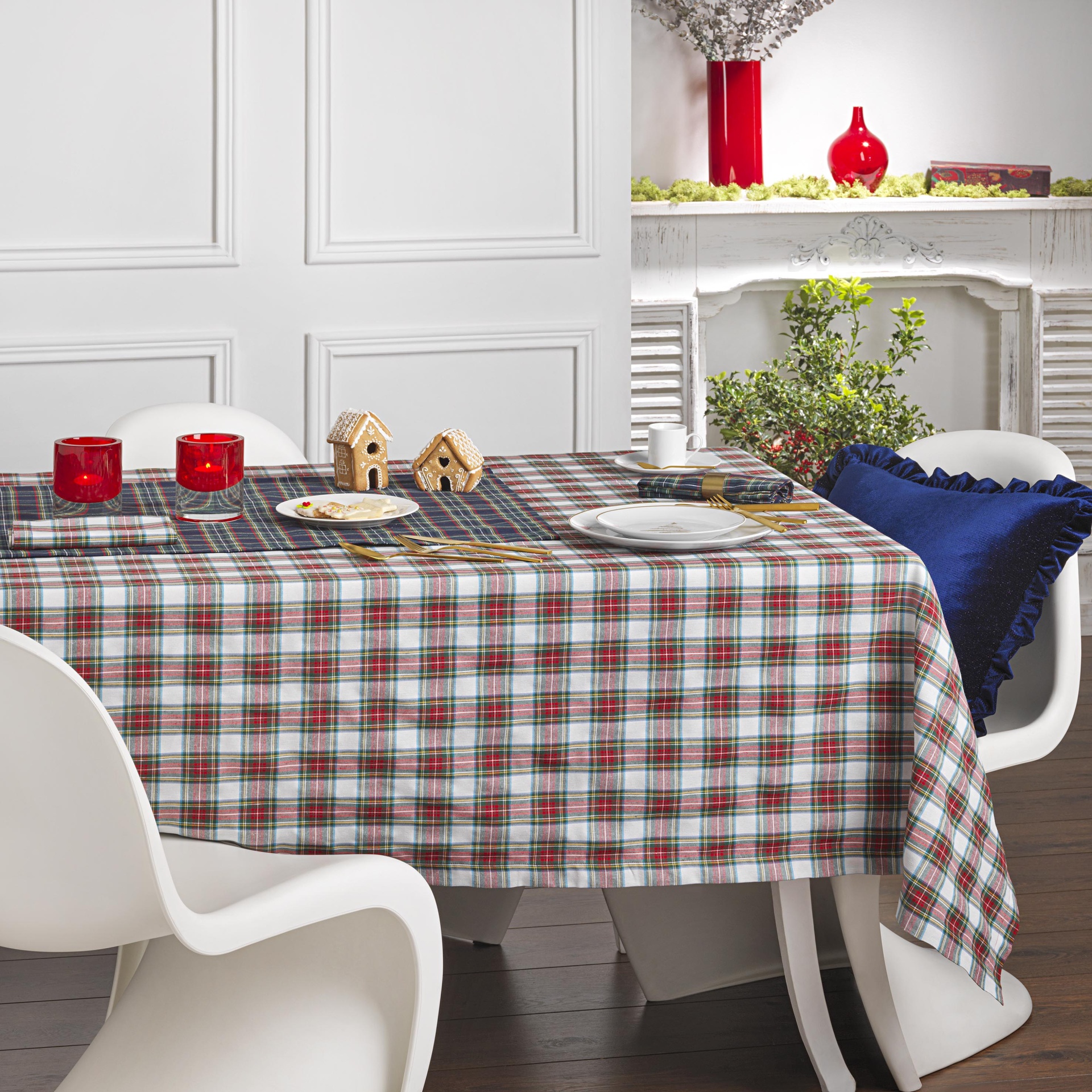 Mesa com toalha xadrez, castiçais, miniaturas de casas, cadeiras brancas e almofada azul num interior com lareira branca e vasos vermelhos