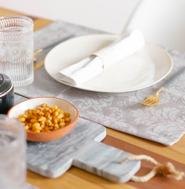 Mesa posta com prato branco, guardanapo branco, copo de vidro, prato de barro com petiscos, garfo dourado e tábua de mármore sobre toalha cinzenta