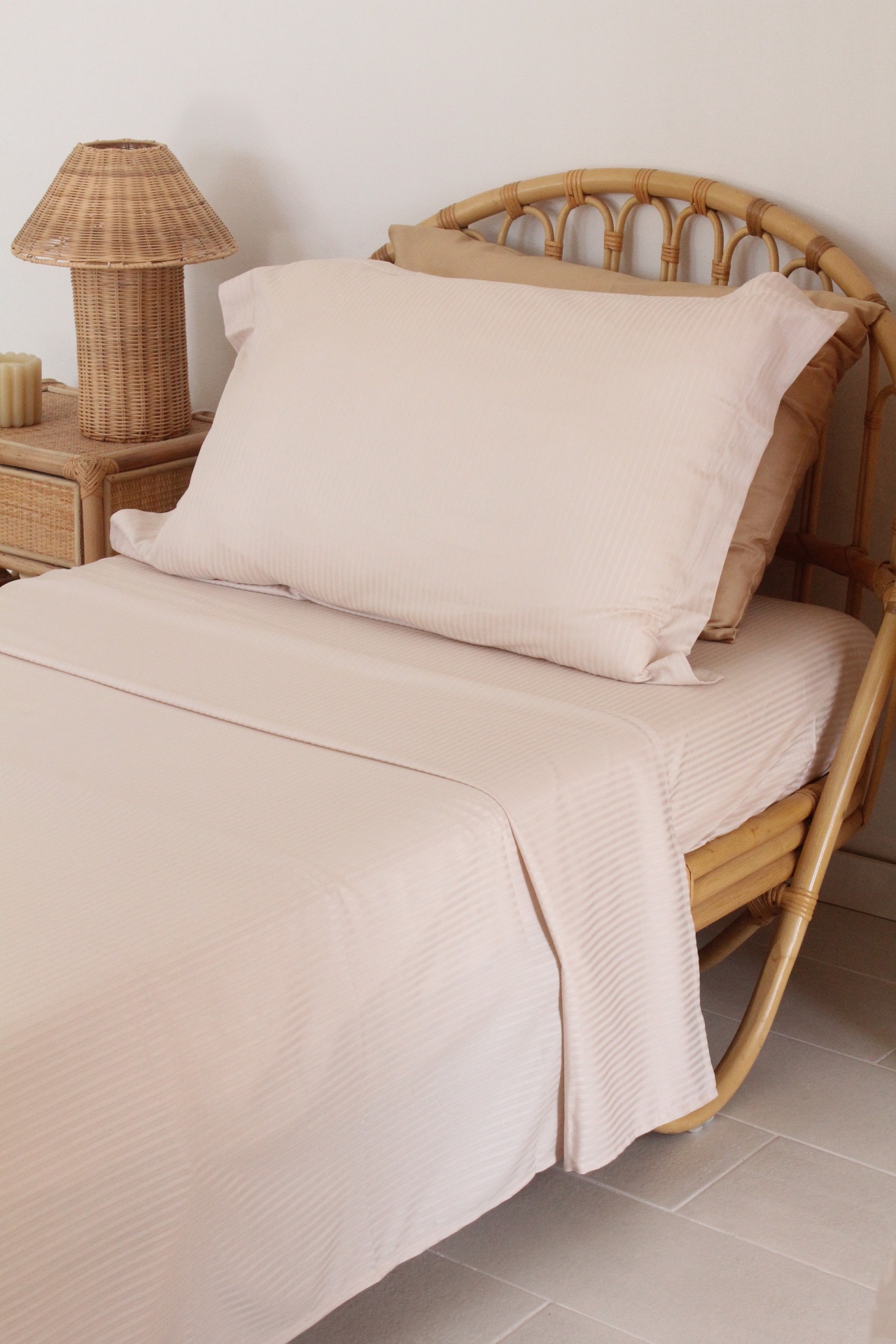 Cama individual com roupa de cama creme com padrão riscas, estrutura e mesa de cabeceira em rattan e candeeiro de vime