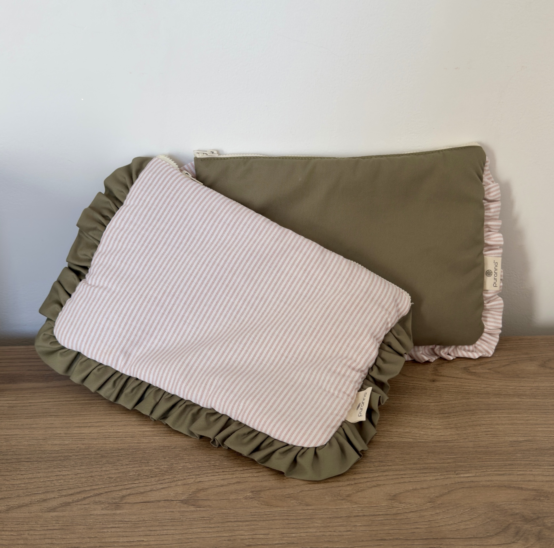 Duas almofadas retangulares com volantes, uma verde-oliva e outra rosa e branca às riscas, com etiquetas Puromar, sobre madeira e parede branca