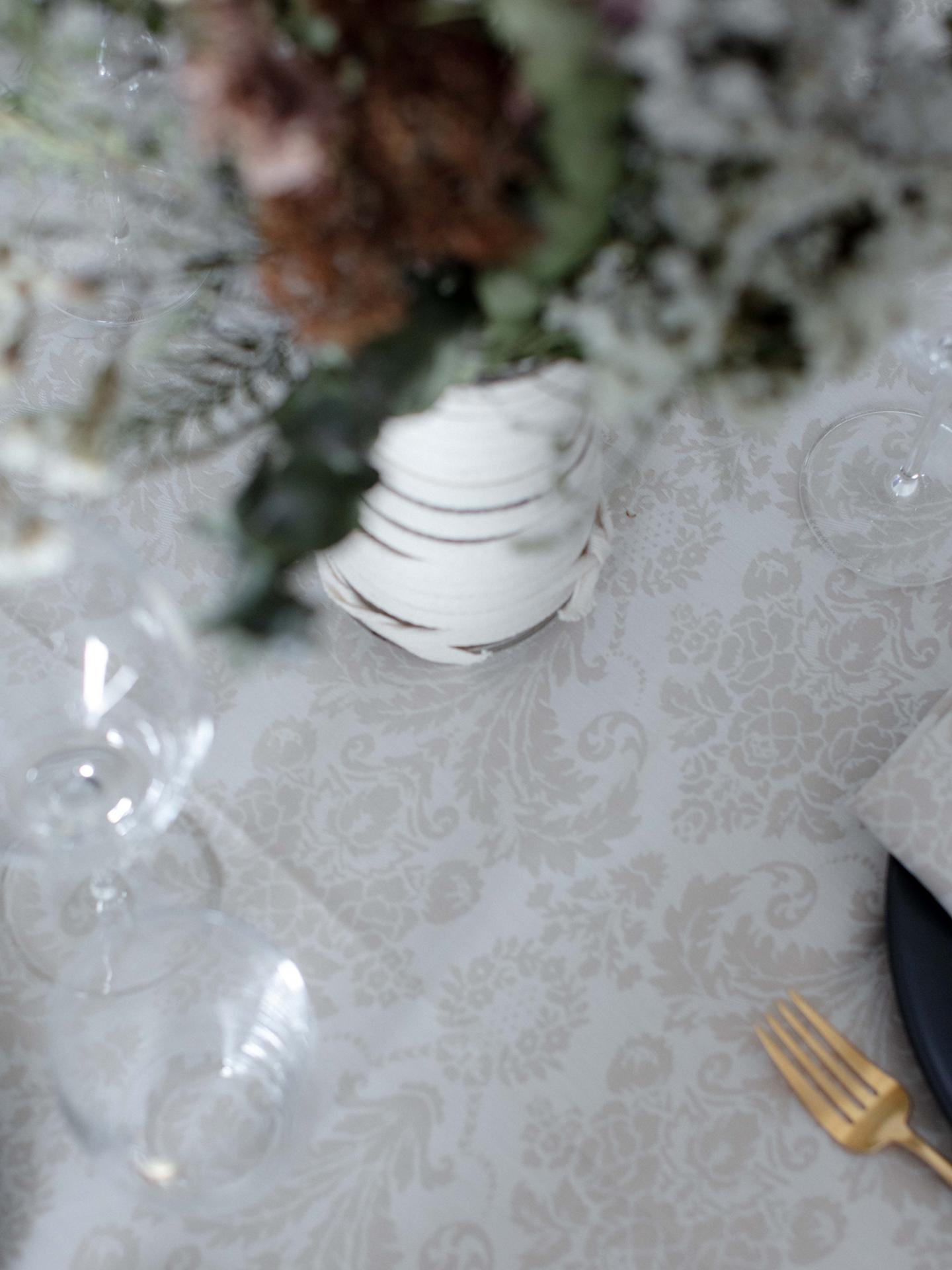 Mesa posta com toalha bege com padrão floral, talher dourado, prato preto, copos de vidro e vaso branco com plantas