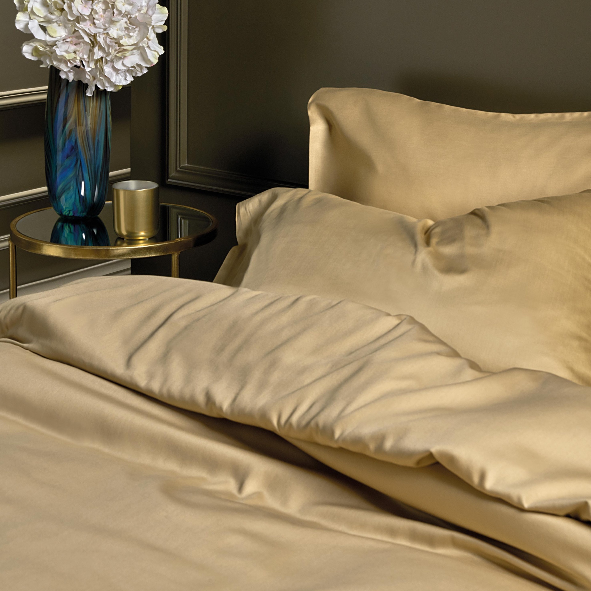 Jogo de cama dourado com almofadas e edredão, ao lado de mesa dourada com vaso azul e flores brancas