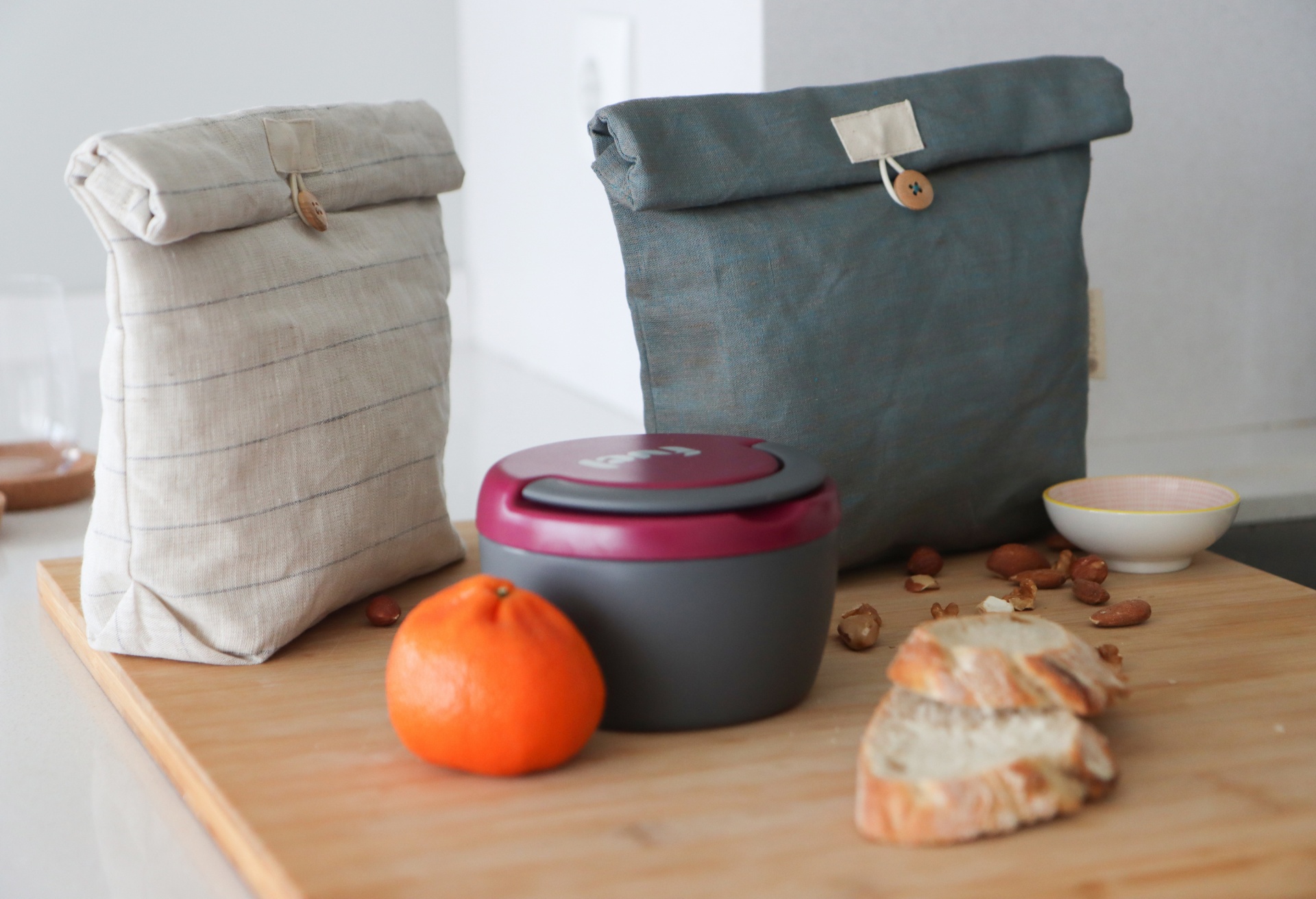 Dois sacos reutilizáveis de tecido com caixa hermética, tangerina, nozes e pão sobre mesa de madeira