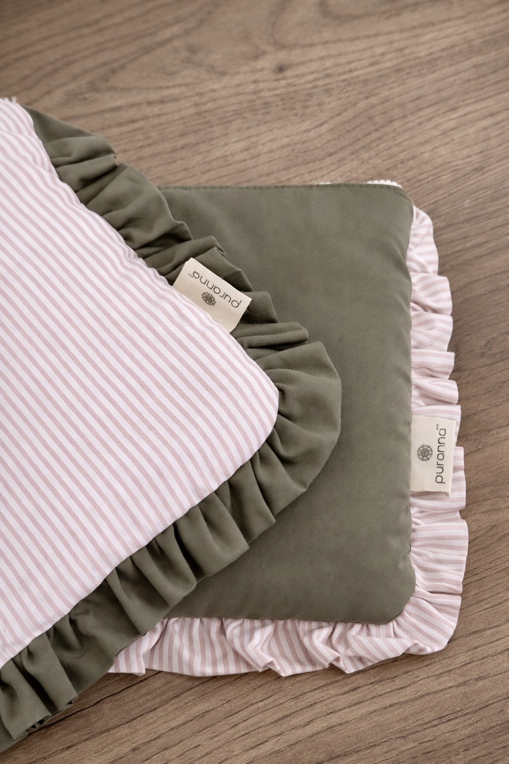 Duas almofadas com folhos, uma verde e outra riscada rosa e branco, com etiquetas puranna, em cima de mesa de madeira.