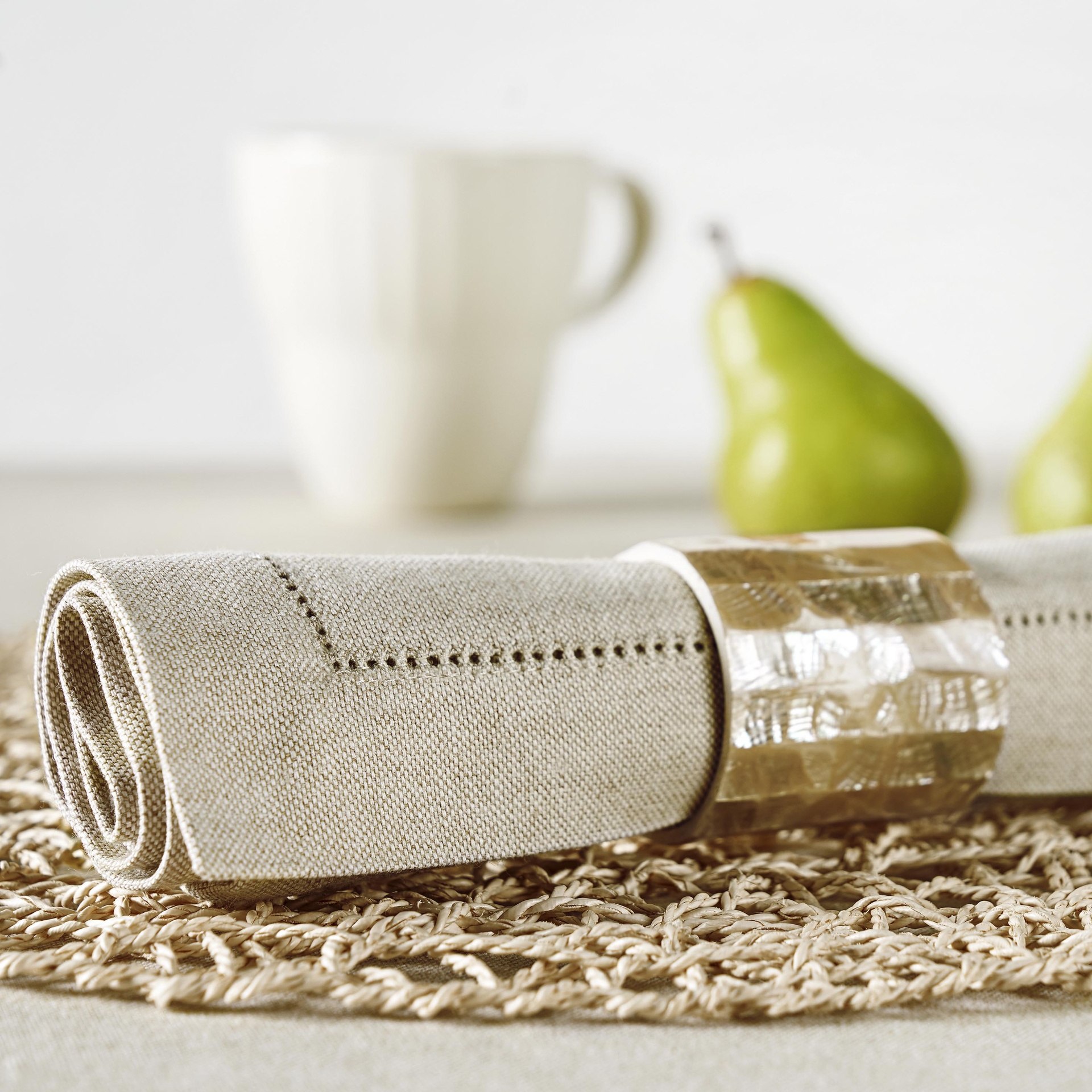 Guardanapo cinzento com argola dourada sobre jogo americano bege, caneca branca e peras ao fundo