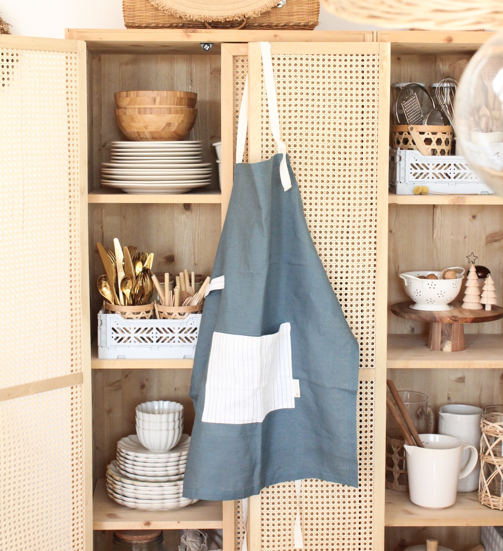 Avental azul com bolso branco pendurado numa porta de armário de cozinha de madeira com pratos e utensílios.