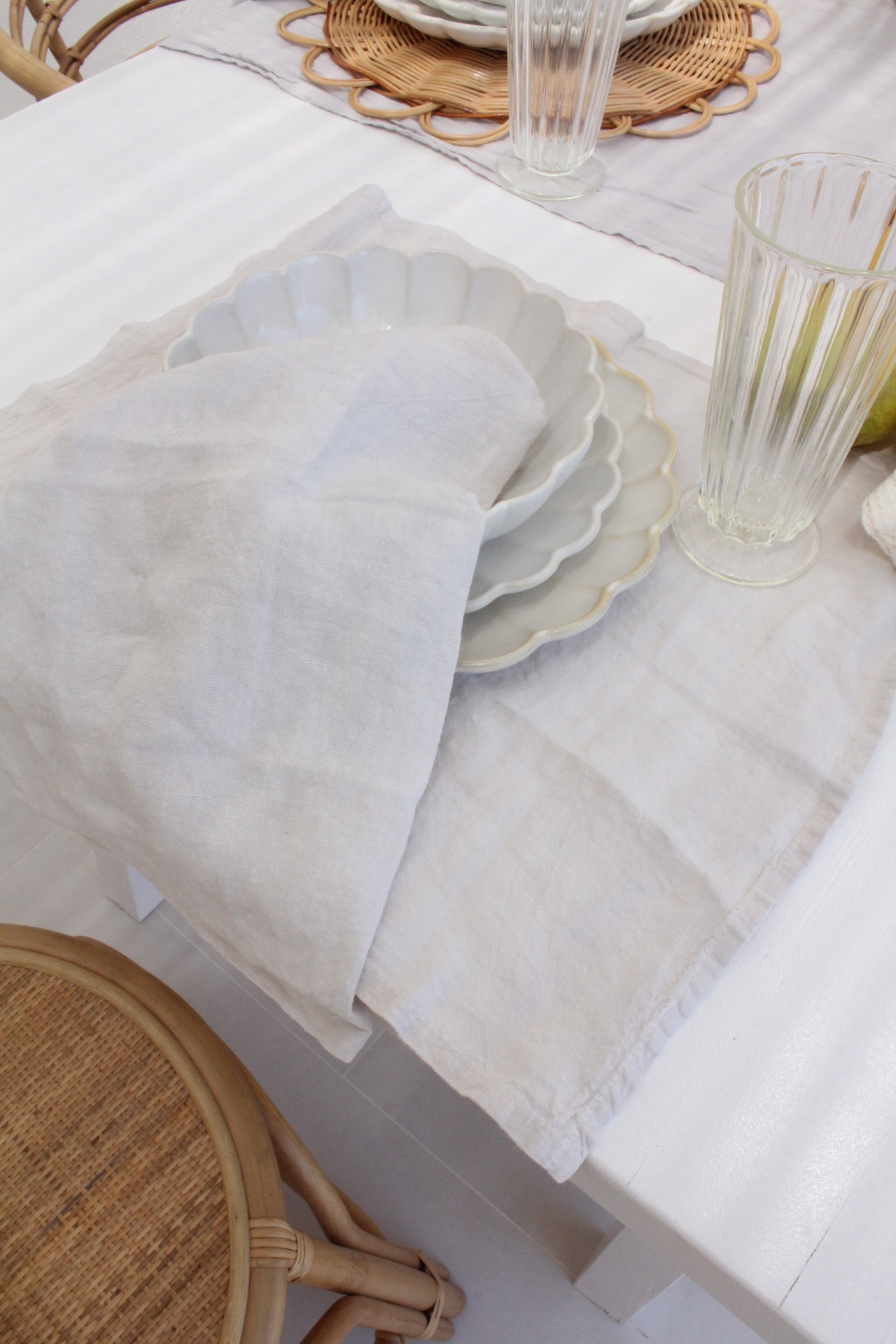 Mesa posta com pratos brancos em camadas e guardanapo bege, copos transparentes, cadeira de madeira e rattan.