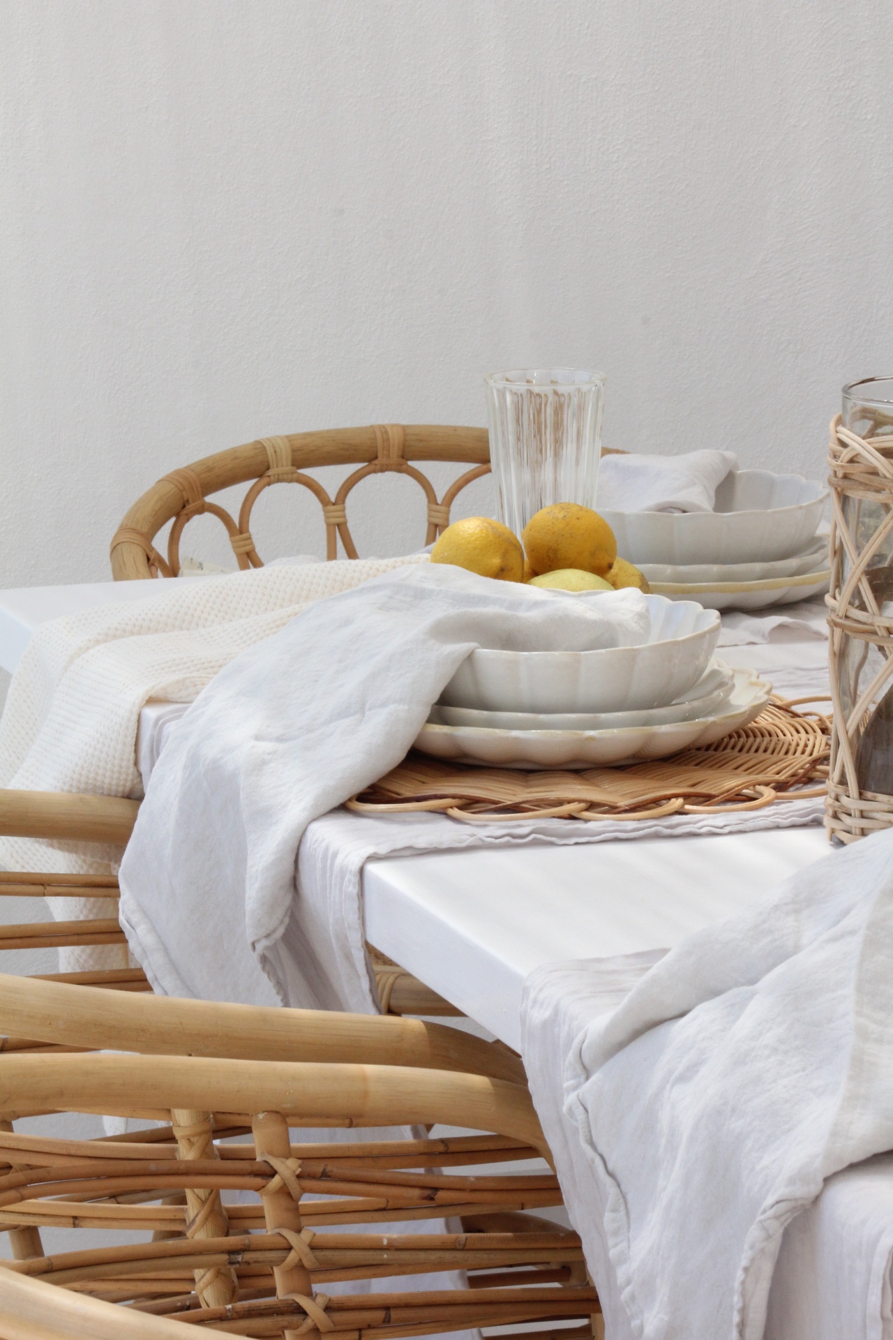 Mesa branca com pratos brancos, limões, copo transparente e cadeiras de rattan