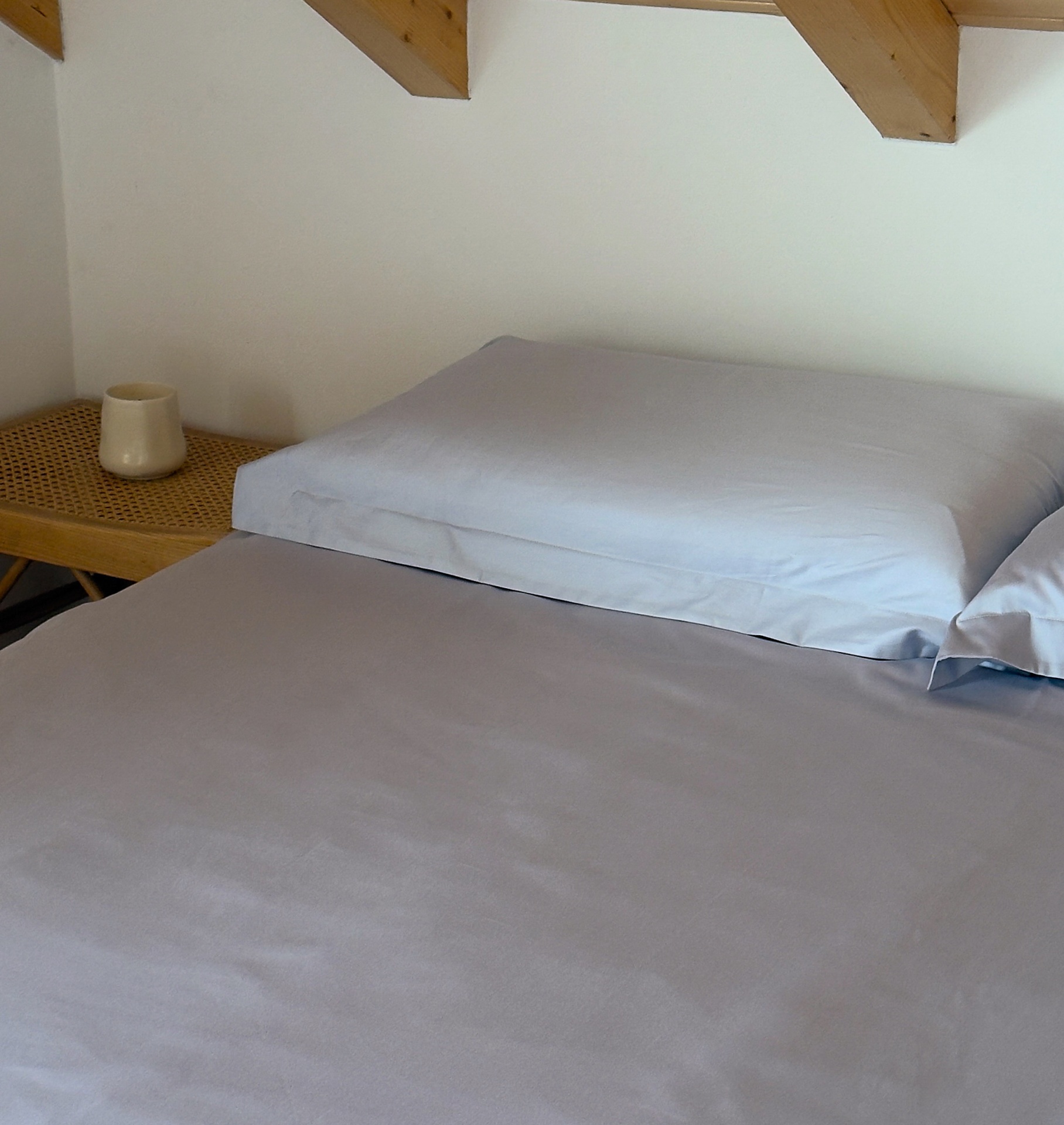 Cama com fronhas cinza sobre colchão e mesa de cabeceira de madeira com caneca creme