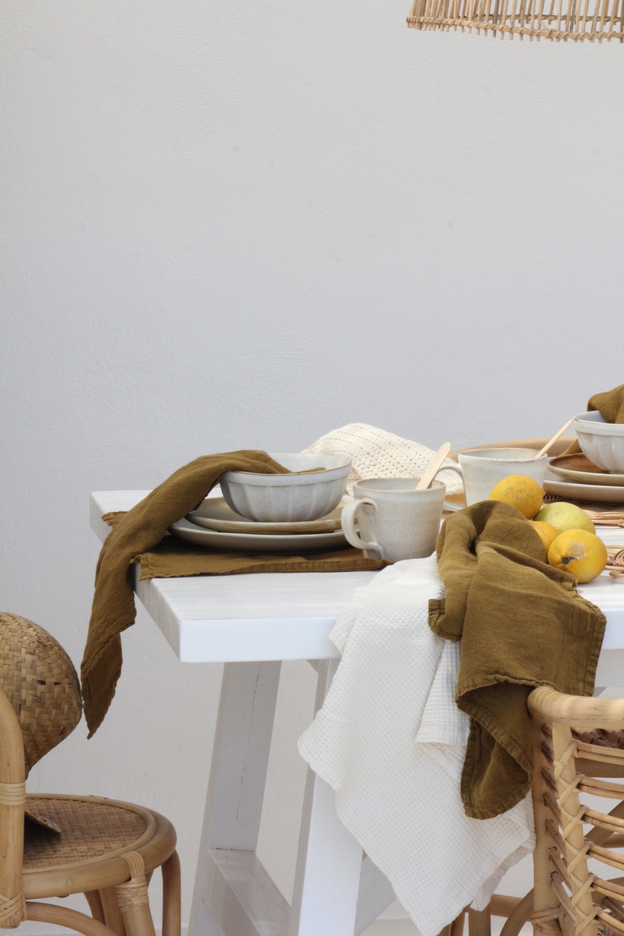 Mesa branca posta com pratos, canecas, talheres de madeira, guardanapos castanhos e brancos, limões, e cadeiras em rattan.