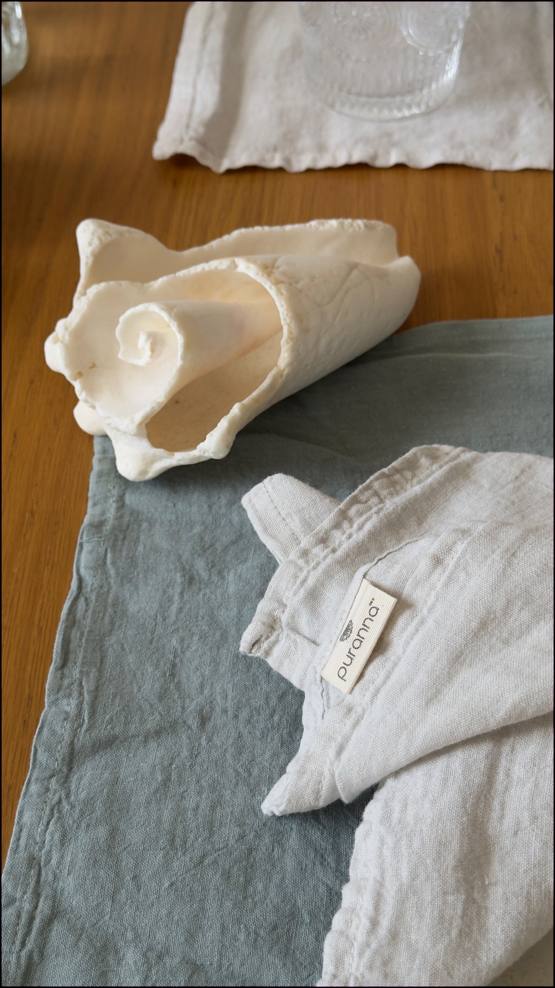 Mesa com guardanapo cinzento-azulado, toalha de linho cinzento clara com etiqueta Puranos, concha decorativa branca e copo transparente