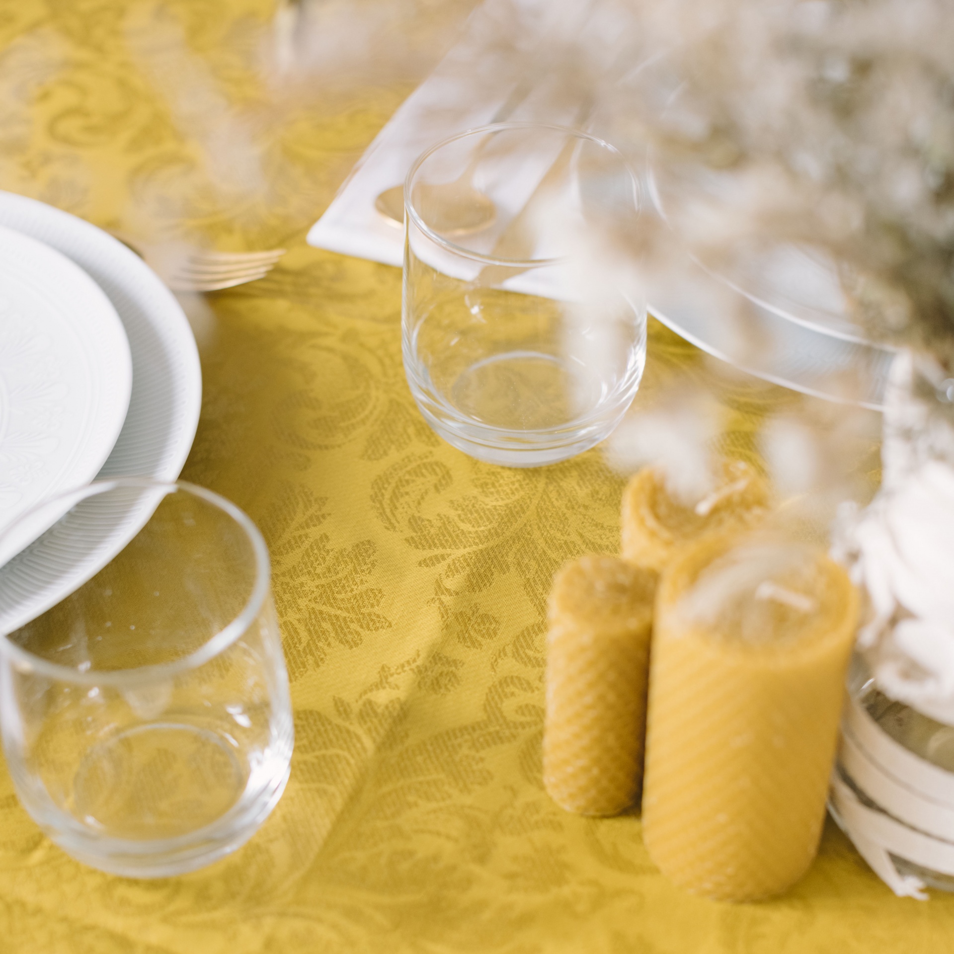 Mesa com toalha amarela, copos de vidro, pratos brancos, velas amarelas e vaso com flores secas.